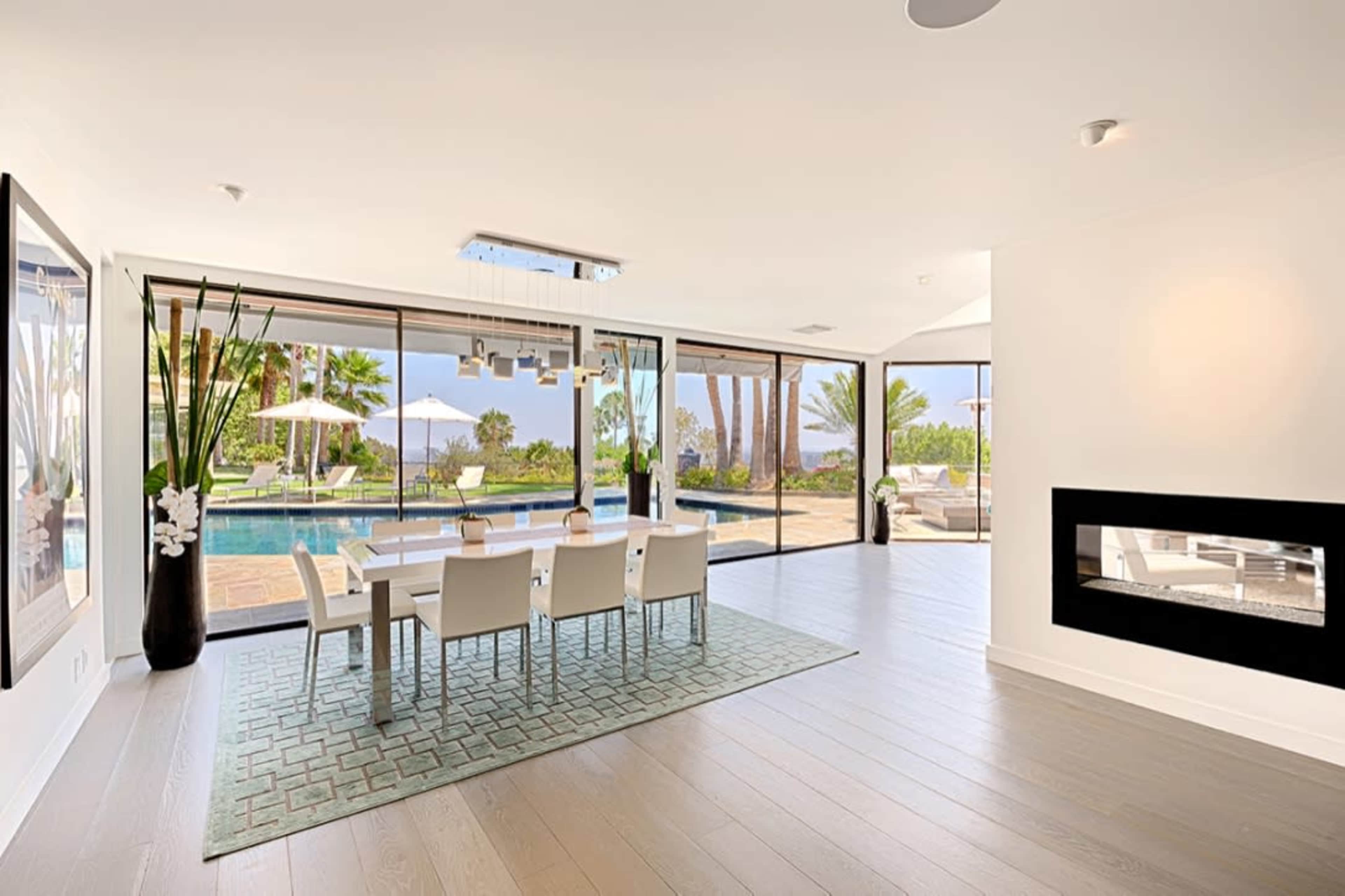 A modern dining area features a large table surrounded by chairs, with large glass doors opening to a poolside view and palm trees outside.