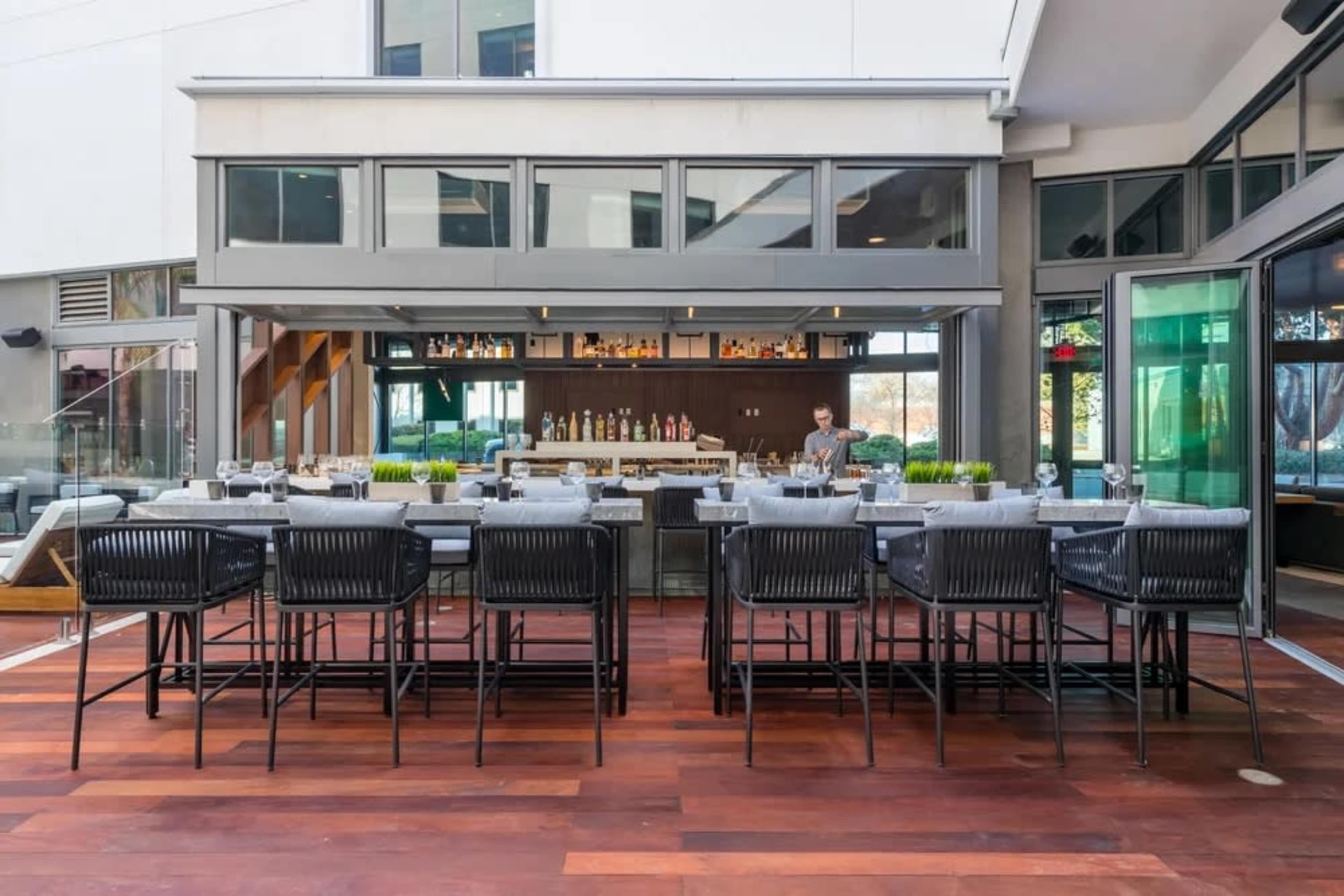 An outdoor bar area features a long counter with high stools and a bartender preparing drinks behind a well-stocked shelf.