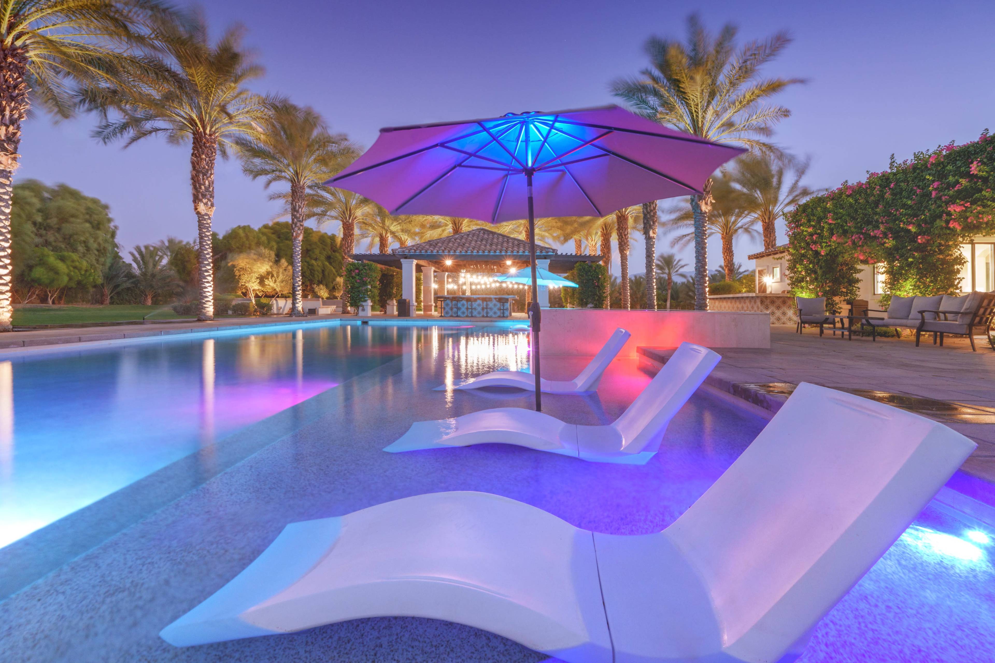 The image shows a swimming pool with lounge chairs and a large umbrella, surrounded by palm trees and illuminated by colorful lights at dusk.