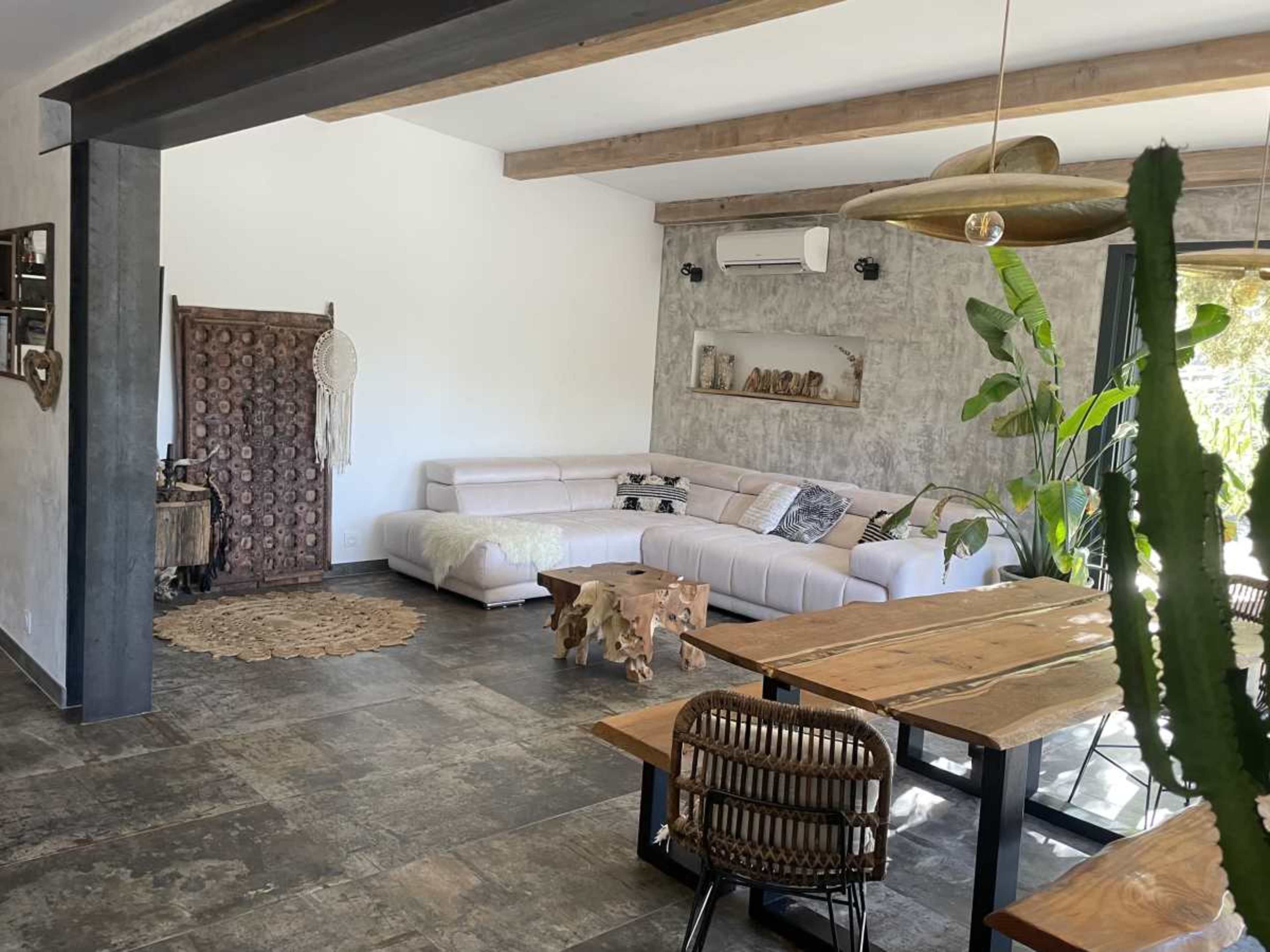The image shows a spacious living area with a white sectional sofa, a wooden dining table, and decorative plants, featuring stone walls and wooden beams.