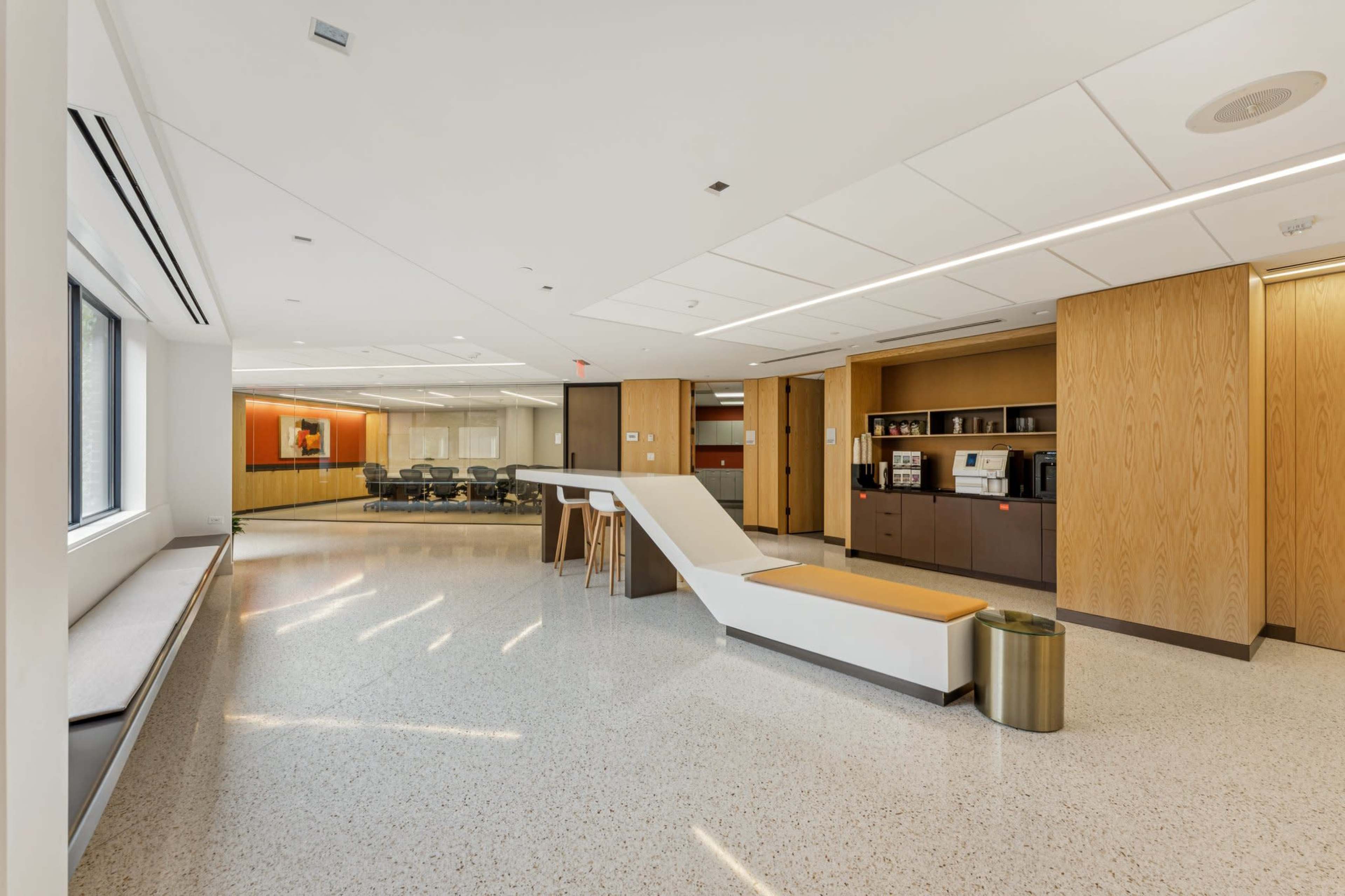 The image shows a modern office interior featuring a sleek seating area, a coffee station, and a conference room in the background.