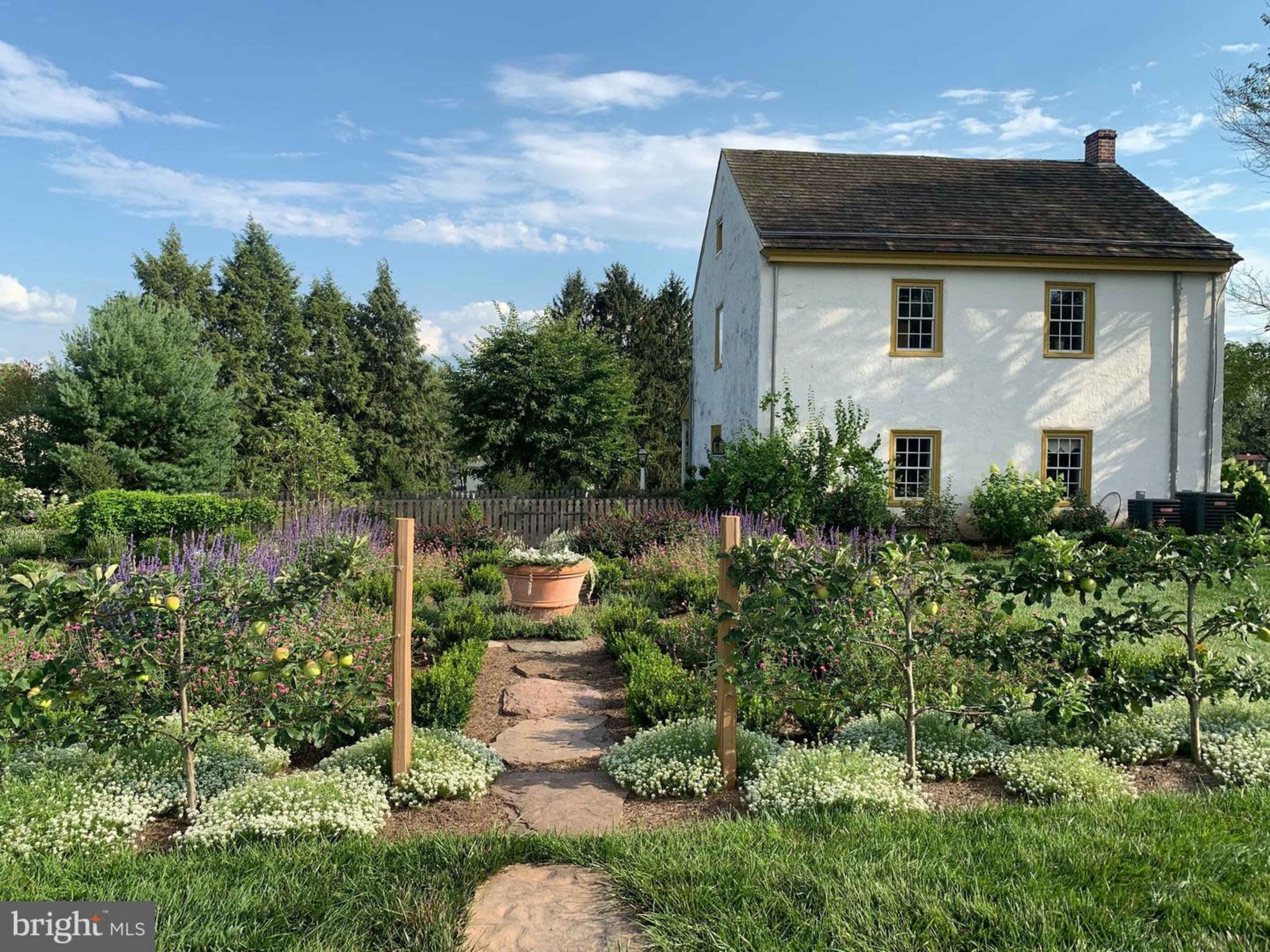 A well-maintained garden featuring flowering plants, shrubs, and ornamental trees surrounds a two-story white house.
