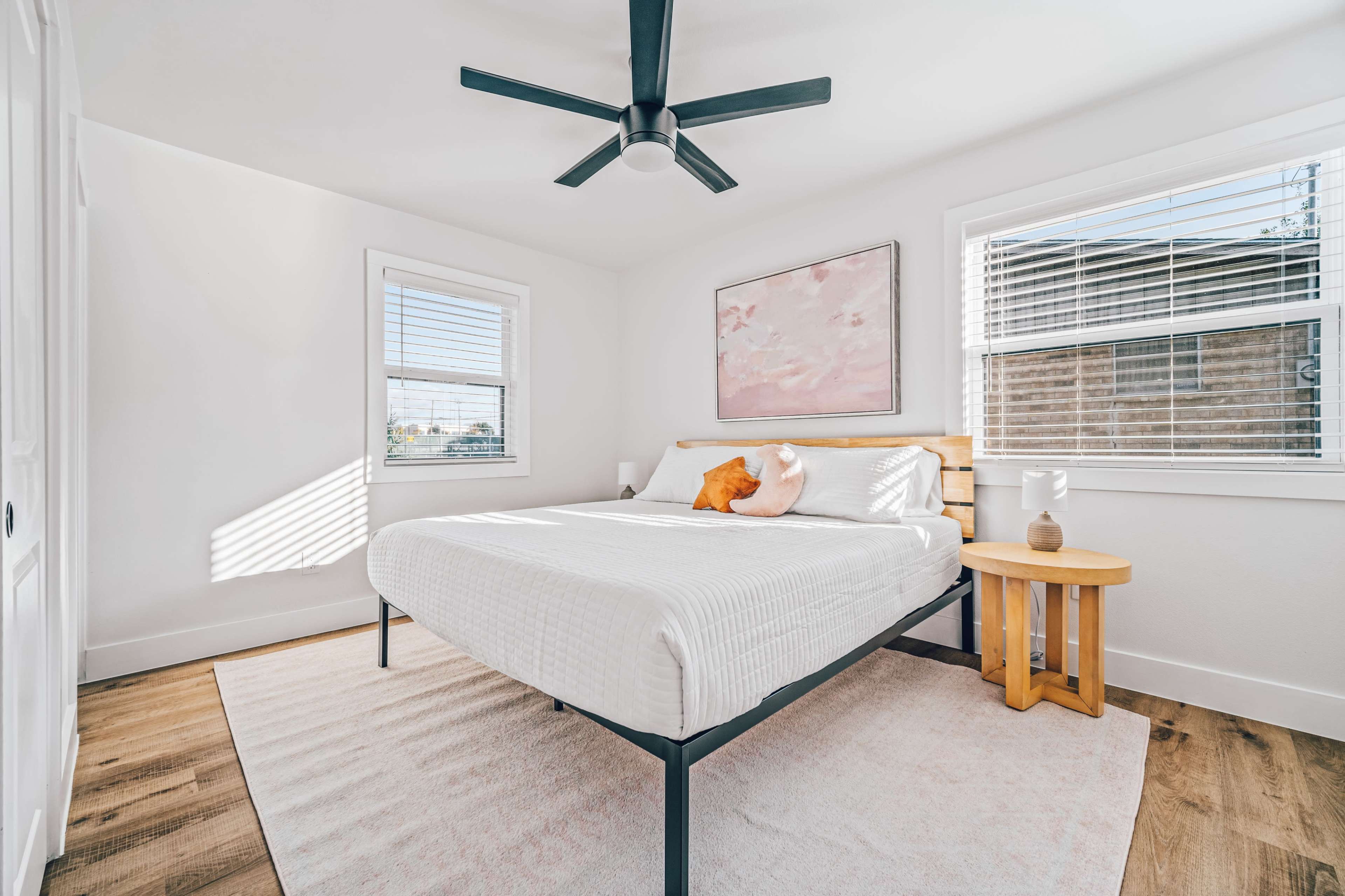 A minimalist bedroom features a bed with a light-colored mattress, a wooden bedside table, and a large window letting in natural light.
