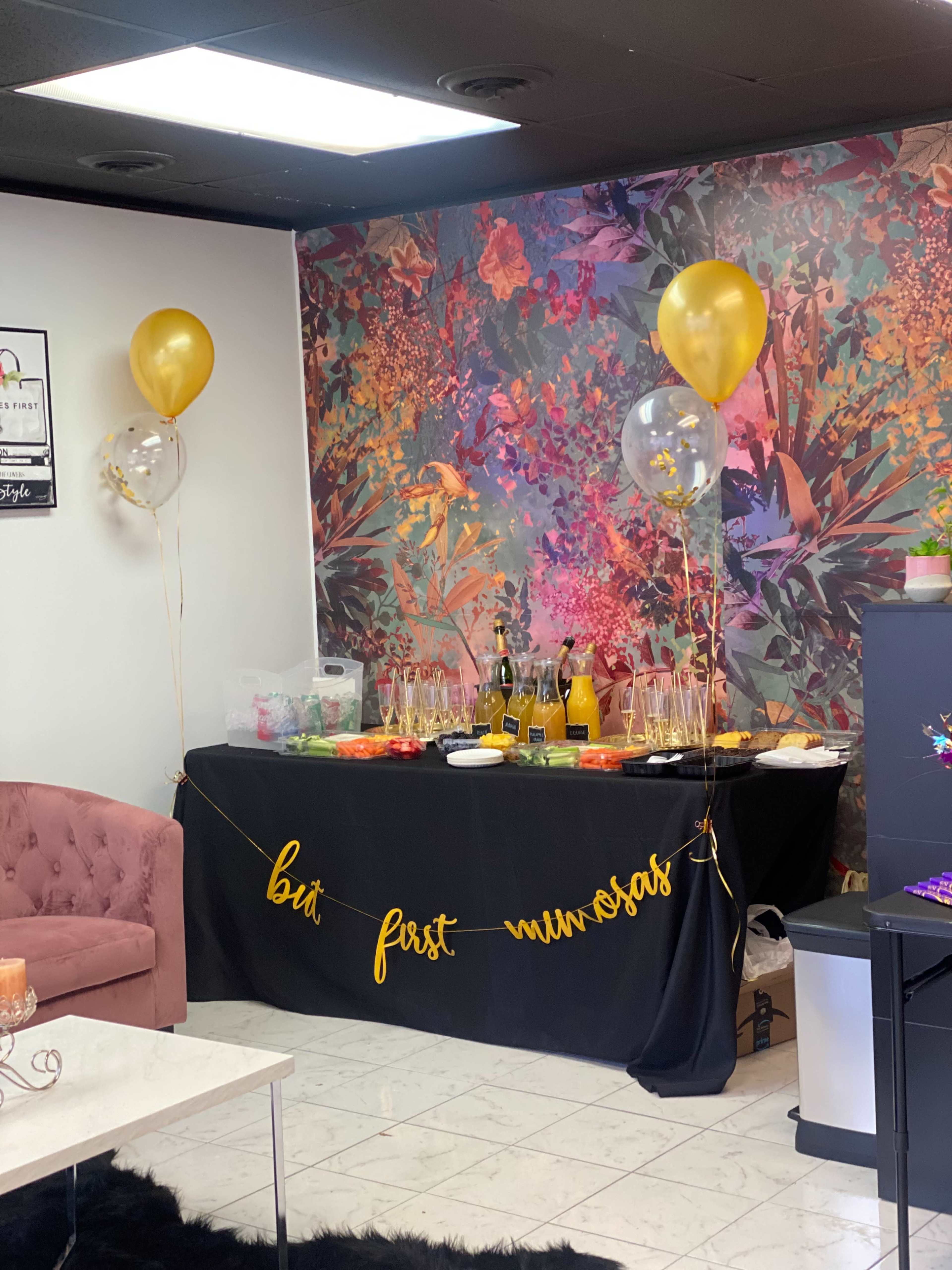 A party setup features a decorated table with snacks, drinks, and balloons against a vibrant floral backdrop.