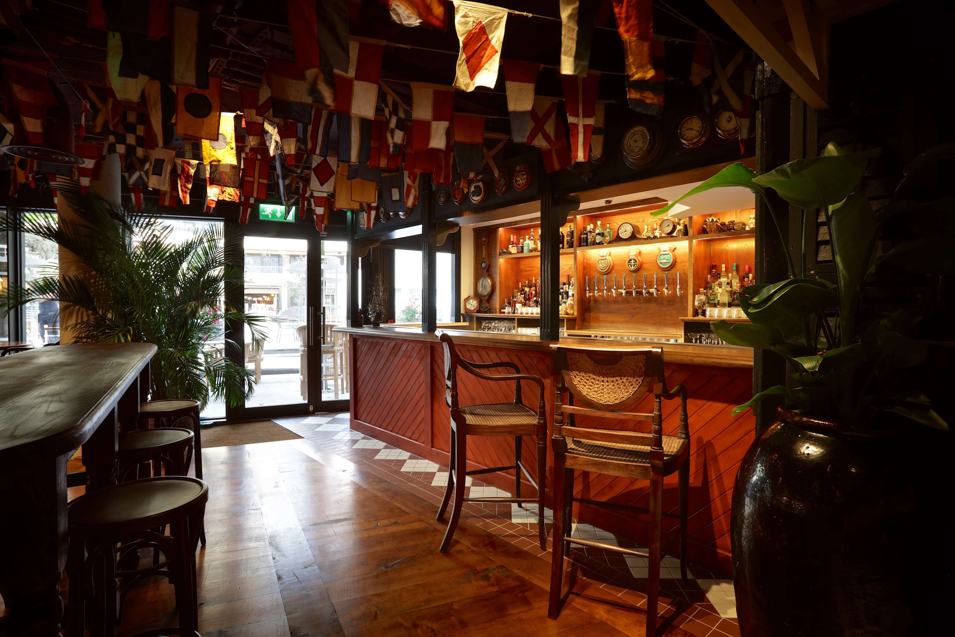 The interior of a bar features a wooden counter, several bar stools, and colorful flags hanging from the ceiling.