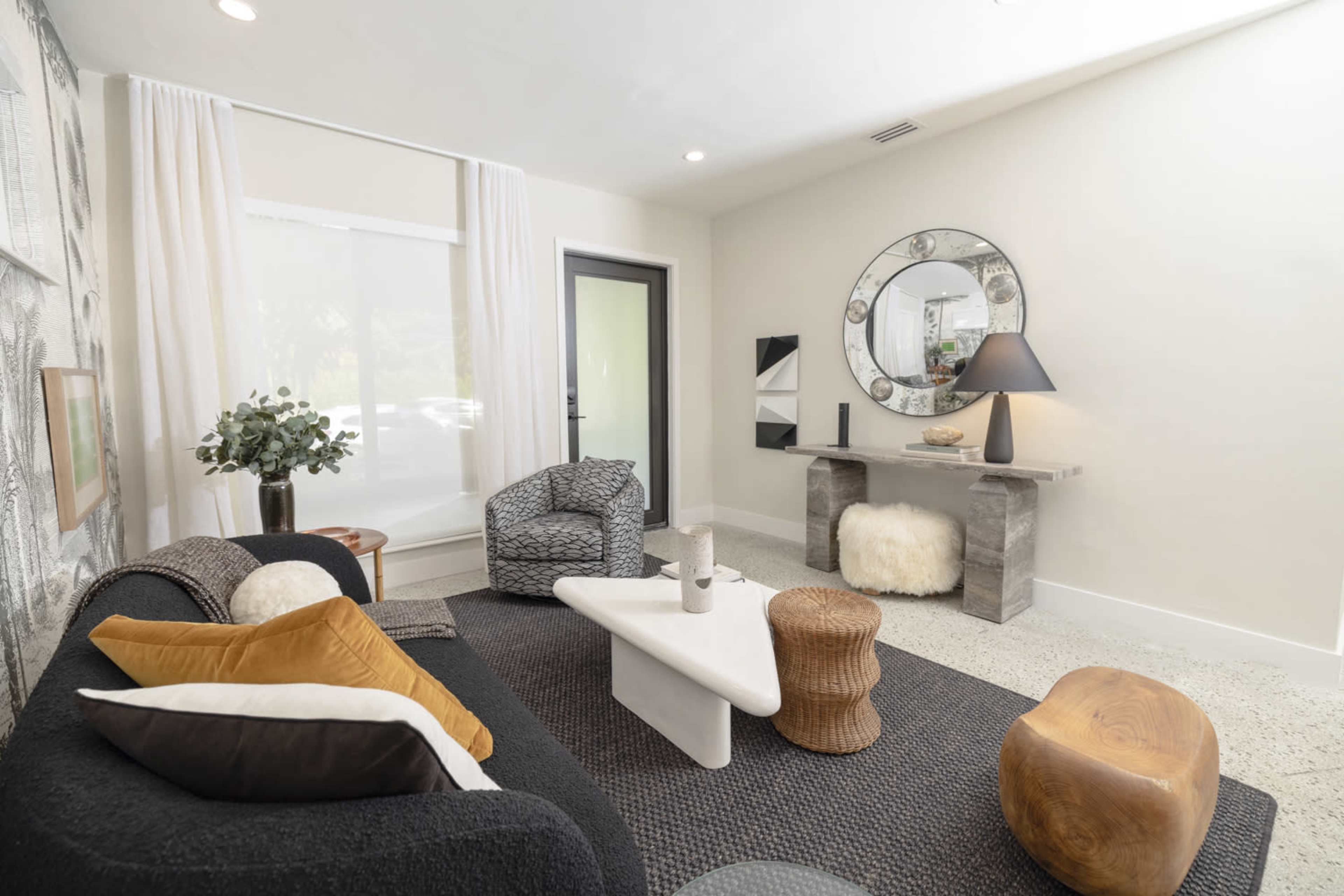 A modern living area with a dark sofa, eclectic seating options, a coffee table, and decorative elements like a round mirror and plants.