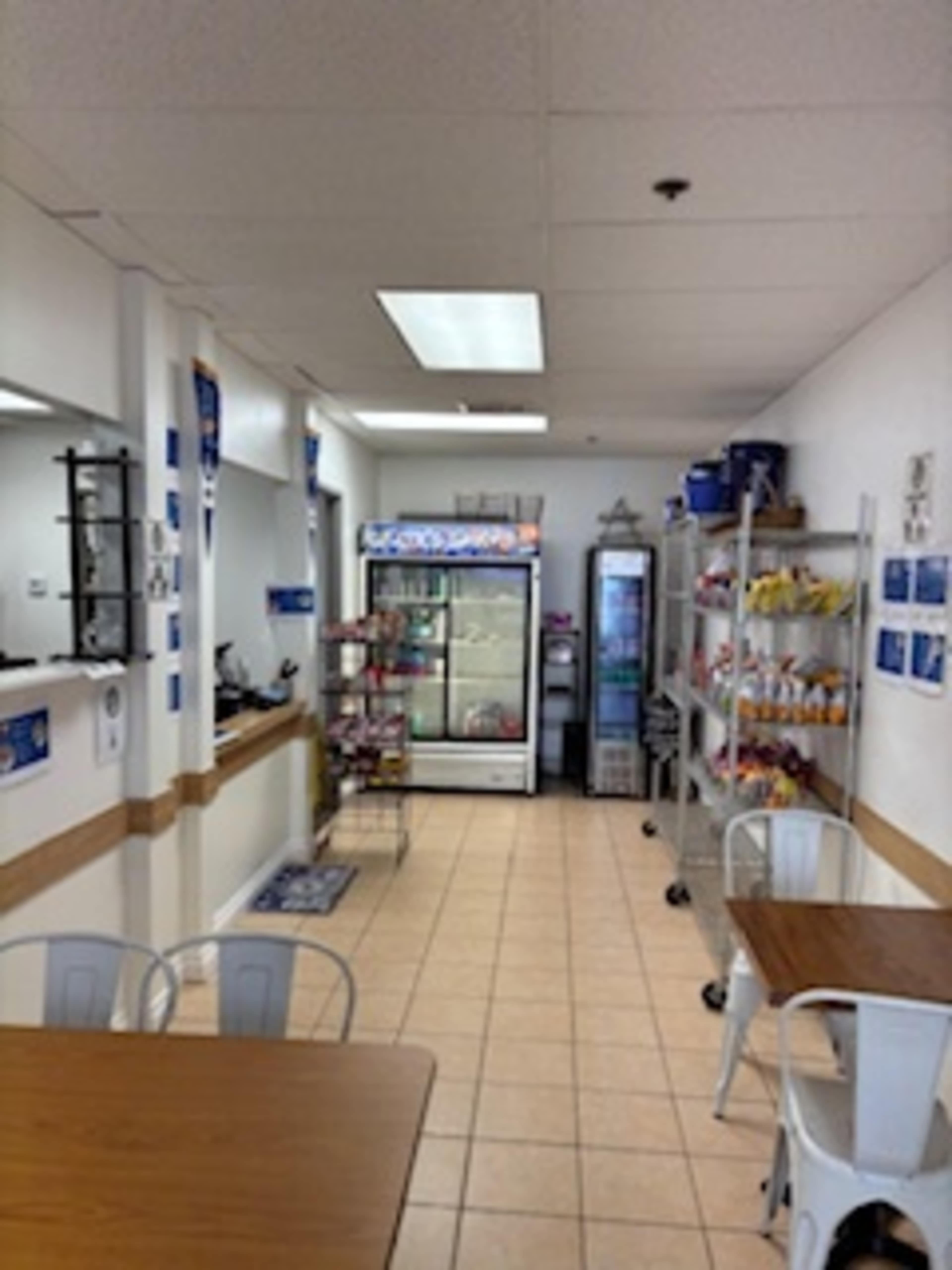 The image shows the interior of a small cafe or convenience store with tables and shelves displaying various products, along with a refrigerator and food cooler.