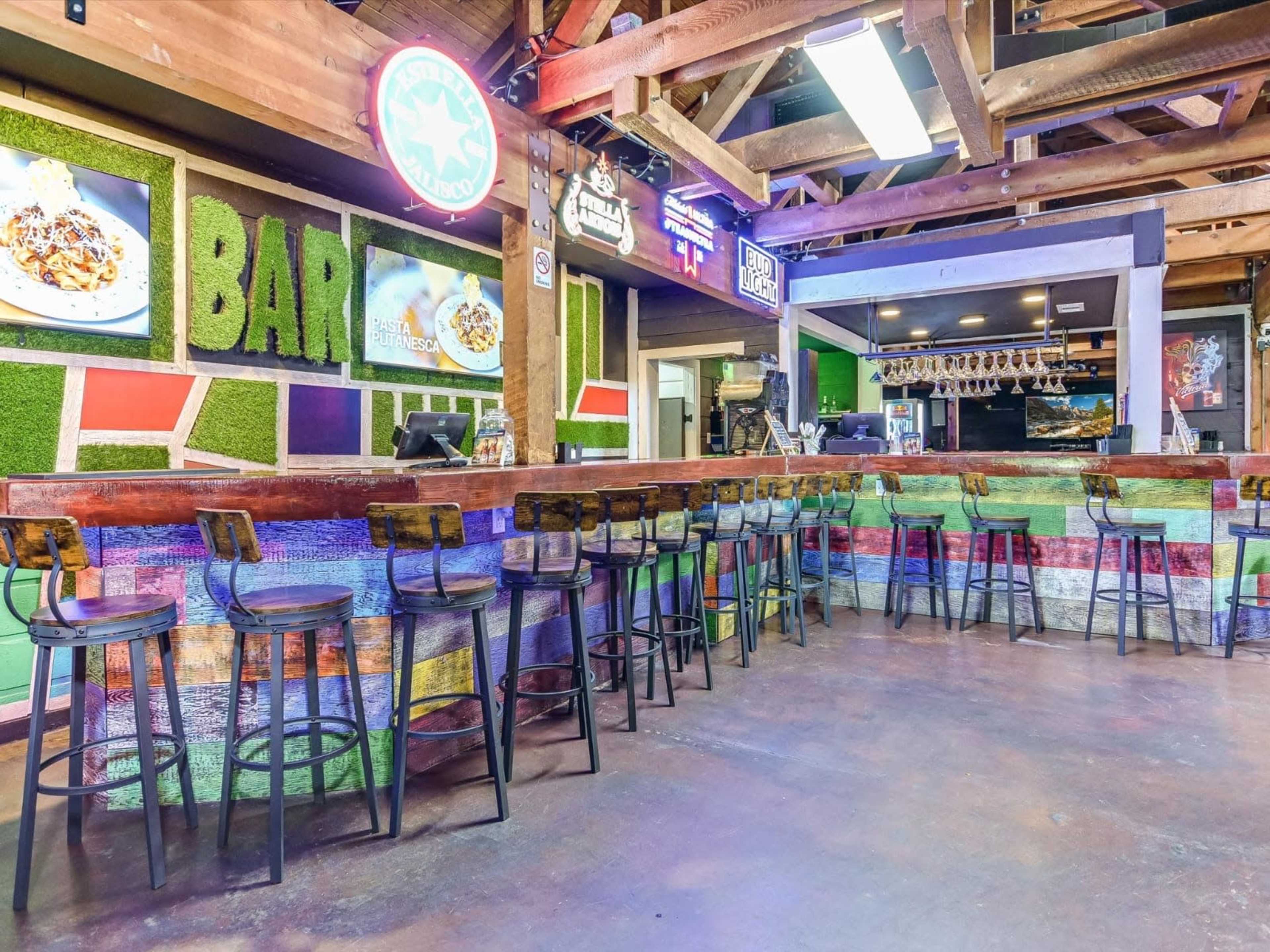 The image shows an empty bar area with colorful decor, featuring wooden stools and a counter, under wooden beams and bright neon signs.