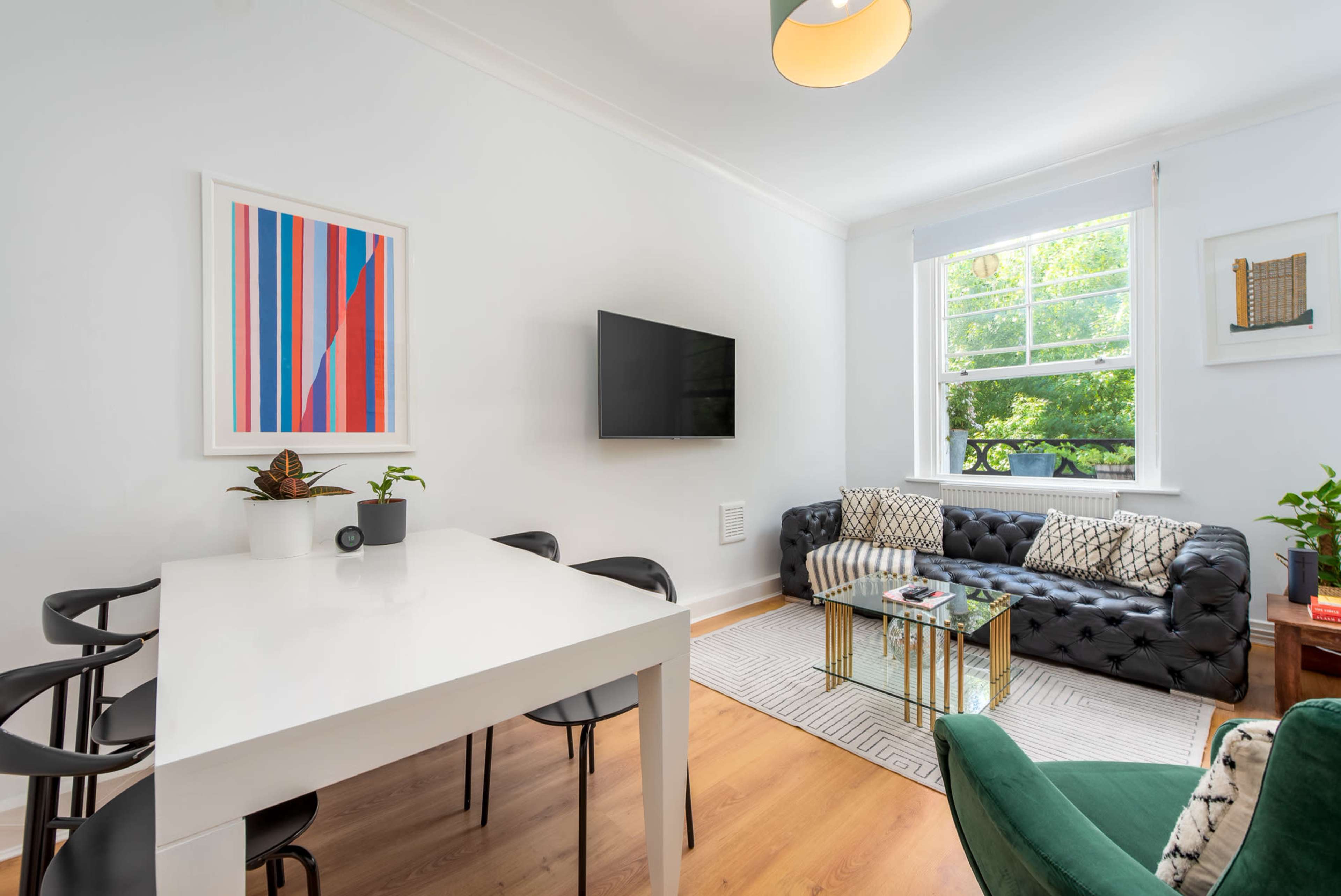A modern living space features a black tufted sofa, a white dining table with black chairs, a wall-mounted television, and large windows letting in natural light.