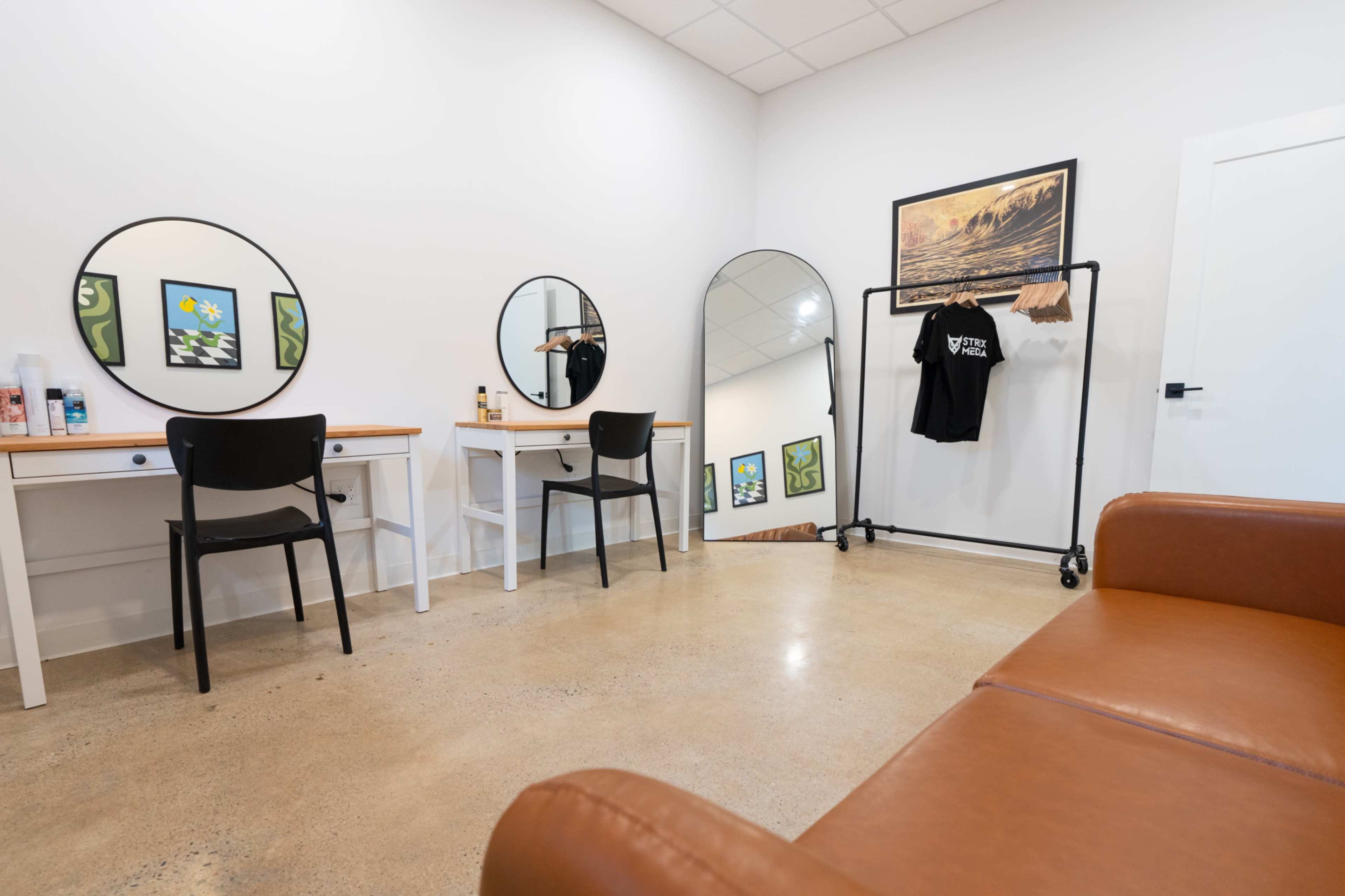 The image shows a modern salon or dressing room with two circular mirrors above white vanities, a black chair at one vanity, a clothing rack with a shirt, and a brown leather couch.