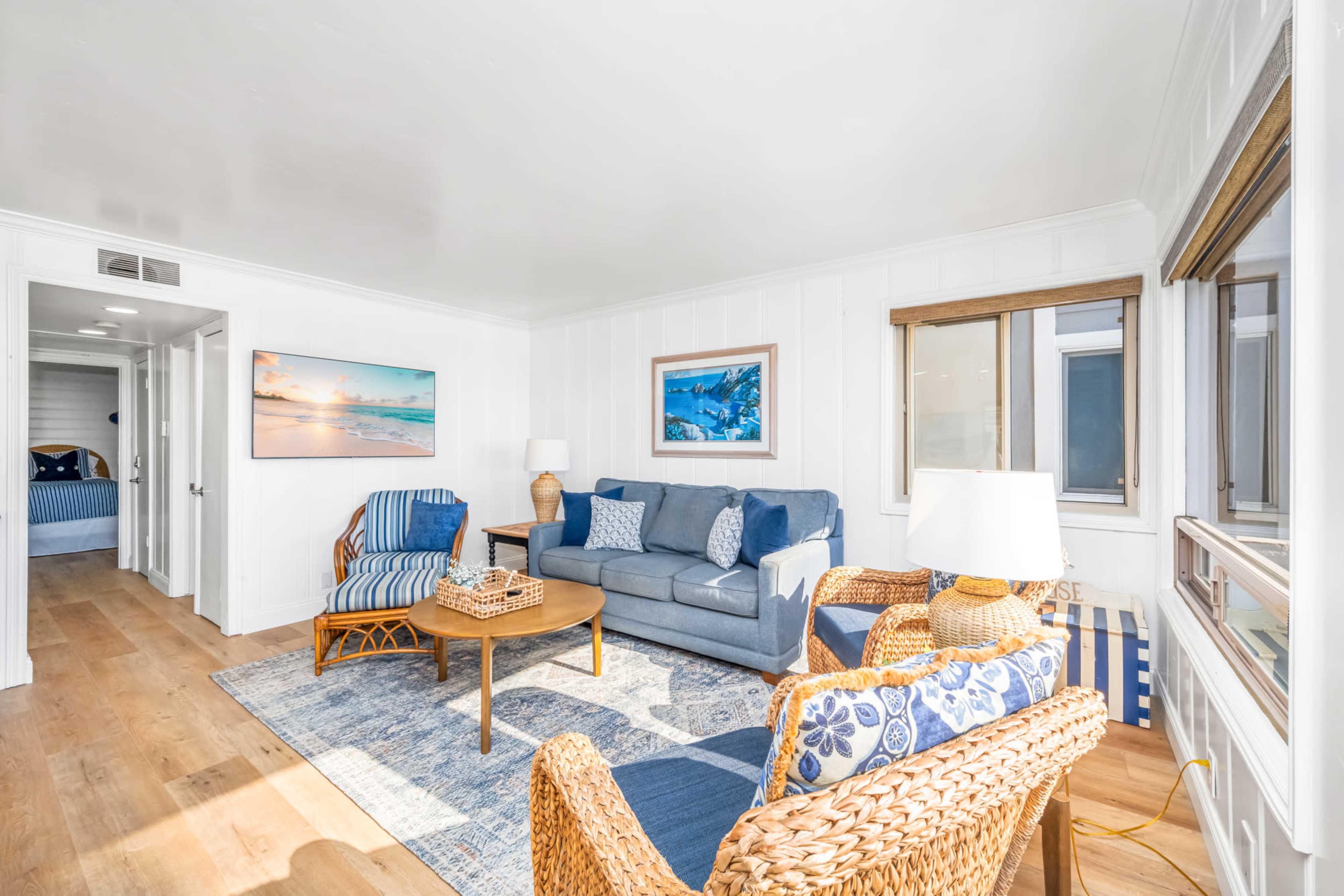A bright living room featuring a blue sofa, wicker chairs, and a coffee table, with a hallway leading to a bedroom visible in the background.