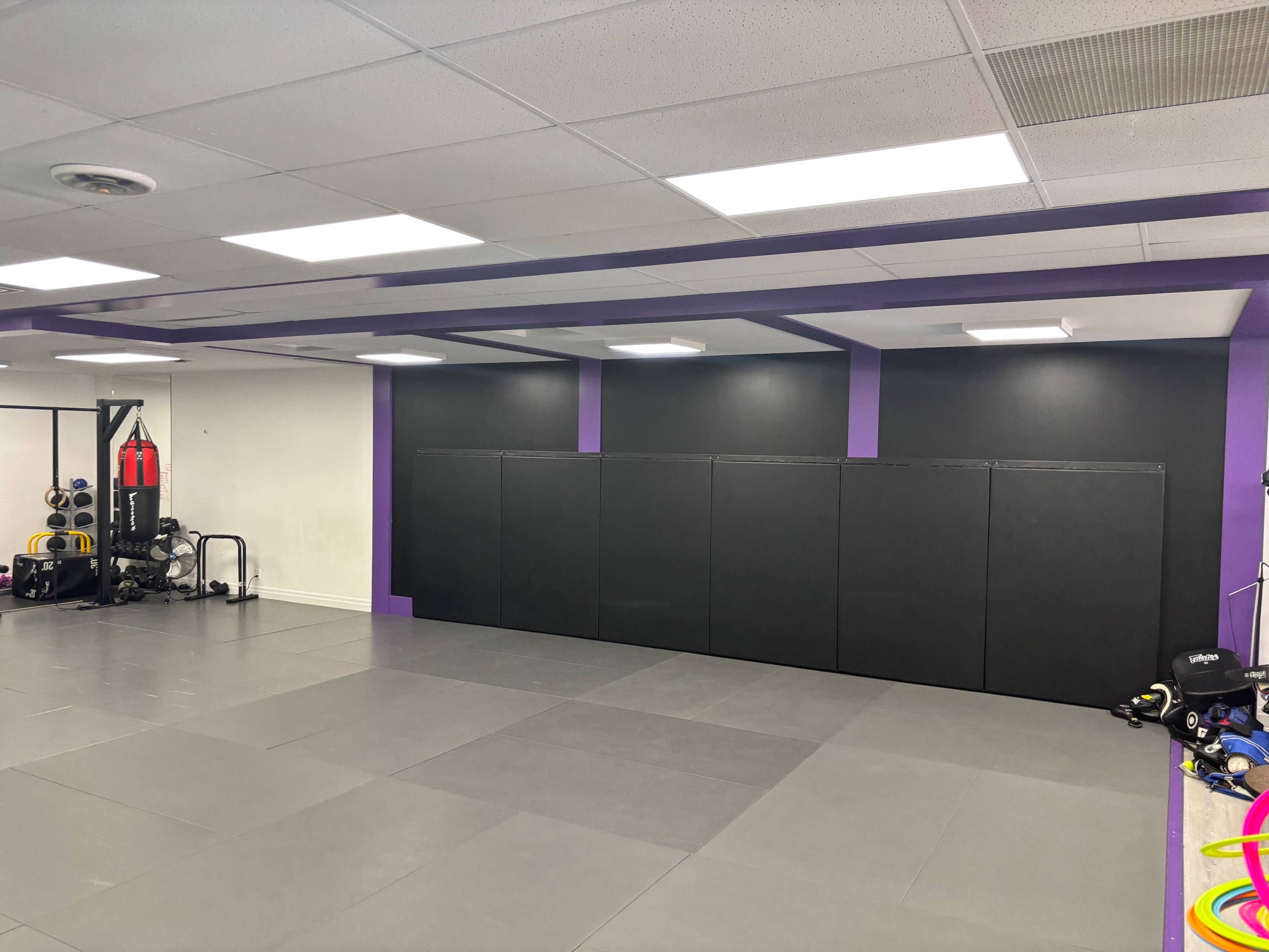 The image shows a gym or training room with gray flooring, black wall pads, and exercise equipment including weights and a punching bag.