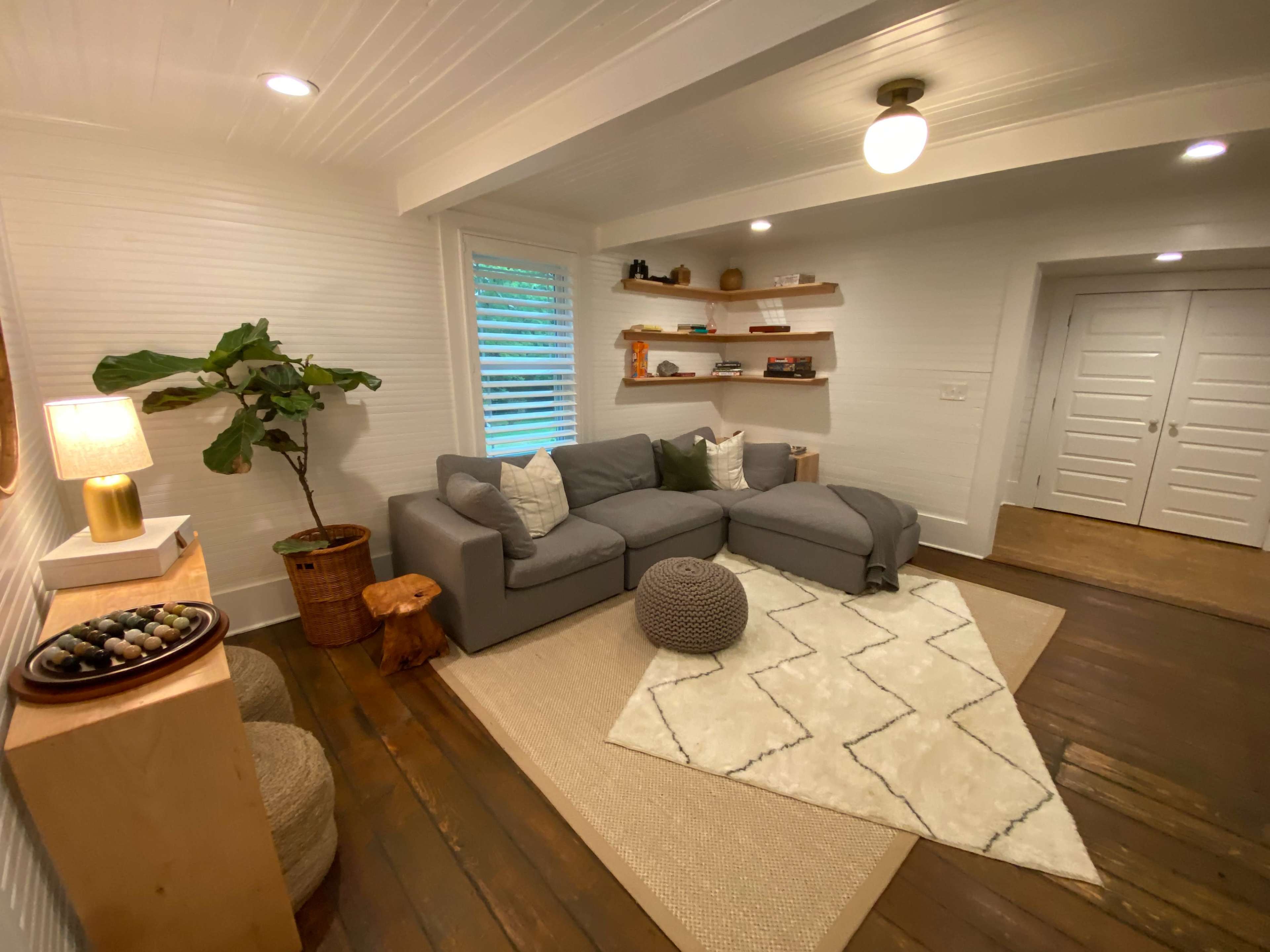 A cozy living room with a gray sectional sofa, a circular pouf, a rug on wooden flooring, and a potted plant beside a lamp and wooden shelves.