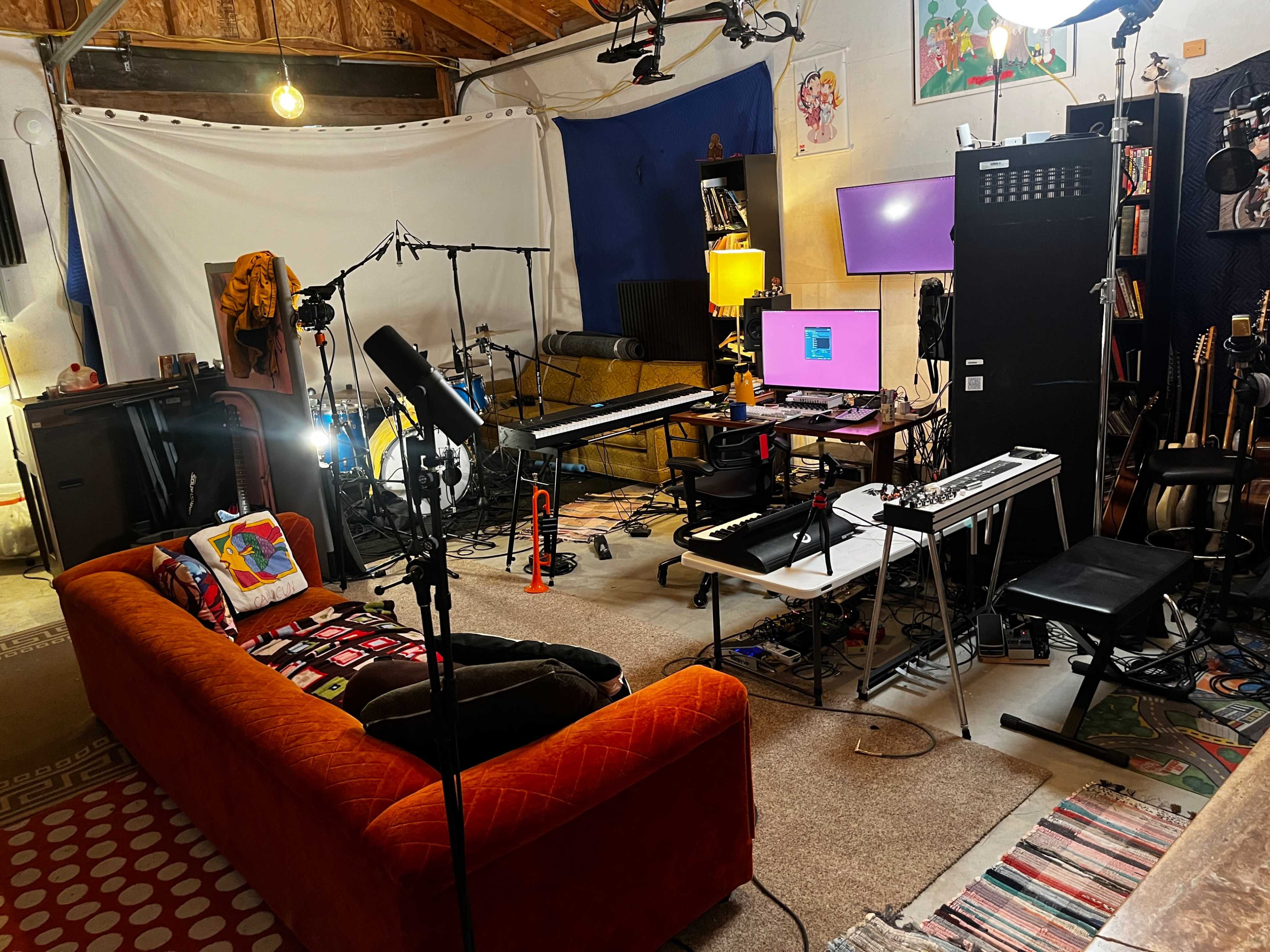 A music studio setup featuring a comfortable orange couch, musical instruments, recording equipment, and various lighting sources.