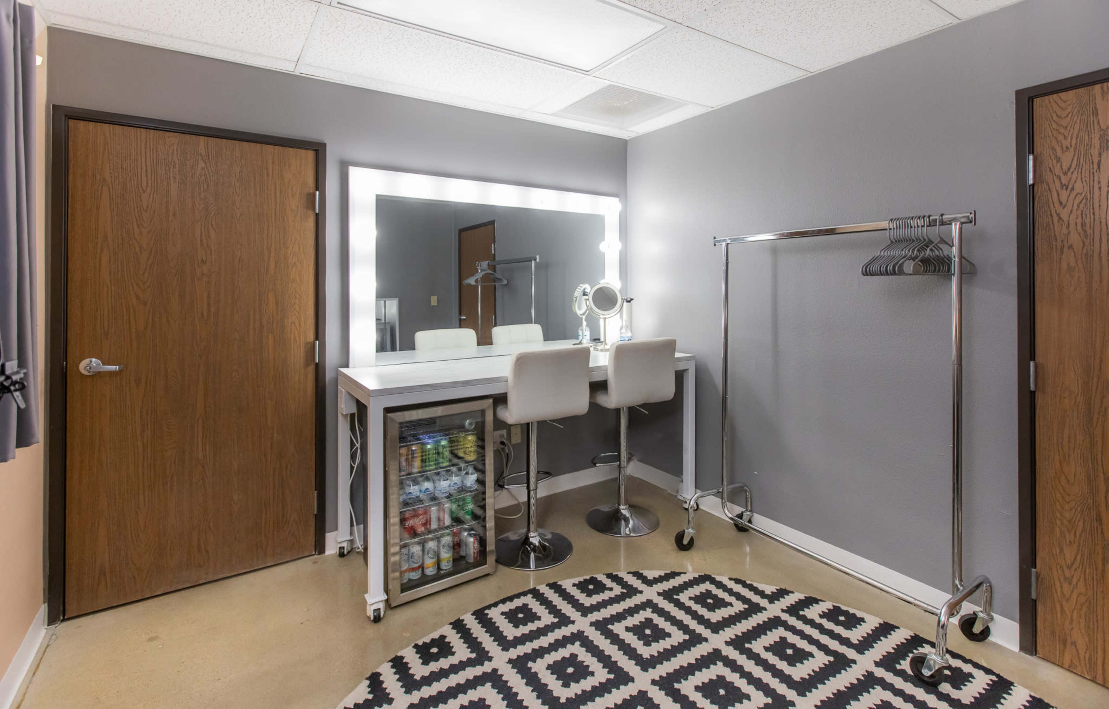 A modern dressing room featuring a makeup vanity with illuminated mirrors, a rack for clothes, and a mini fridge on a patterned rug.