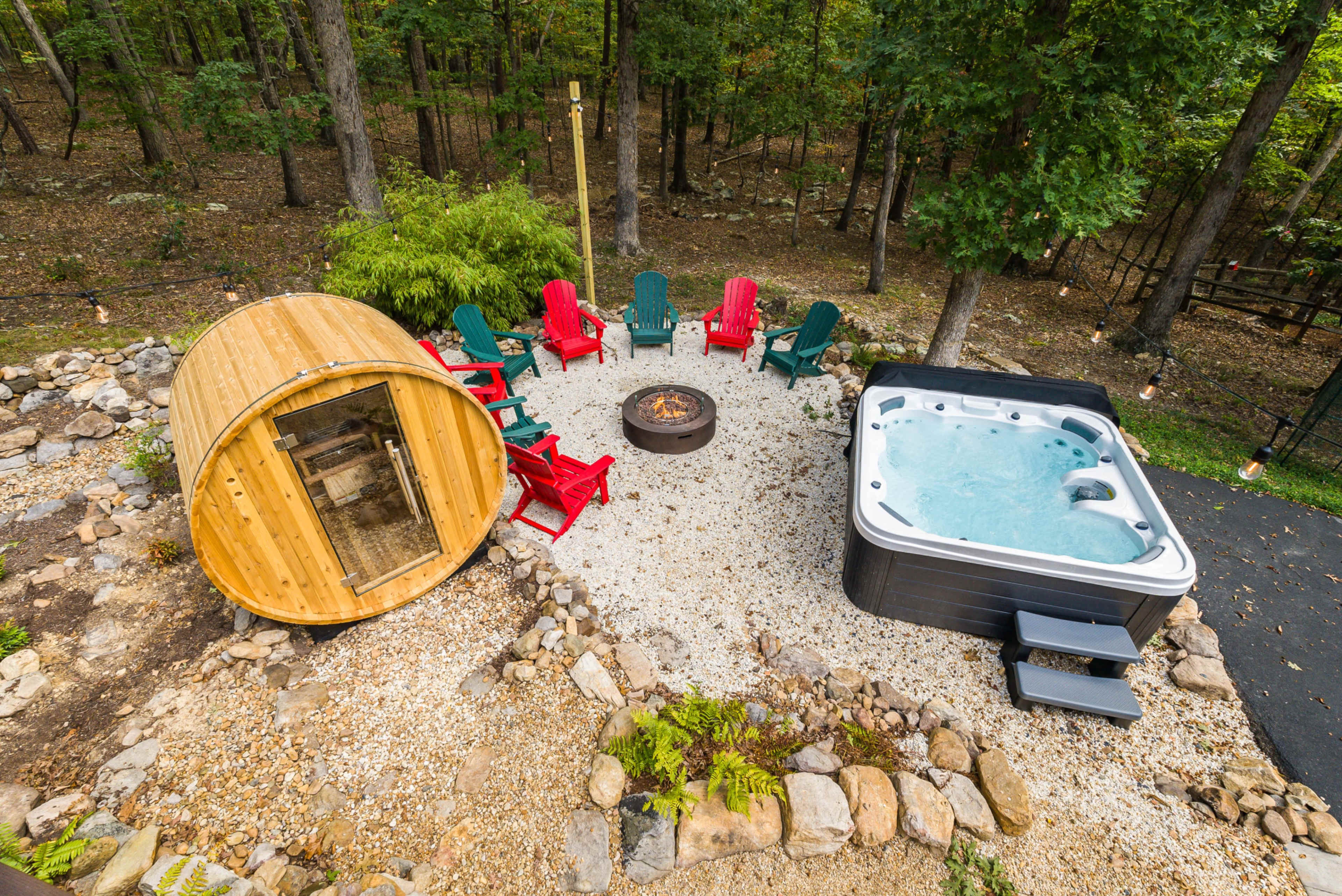The image shows a relaxing outdoor area featuring a wooden sauna, a hot tub, a fire pit surrounded by seating, and a wooded backdrop.