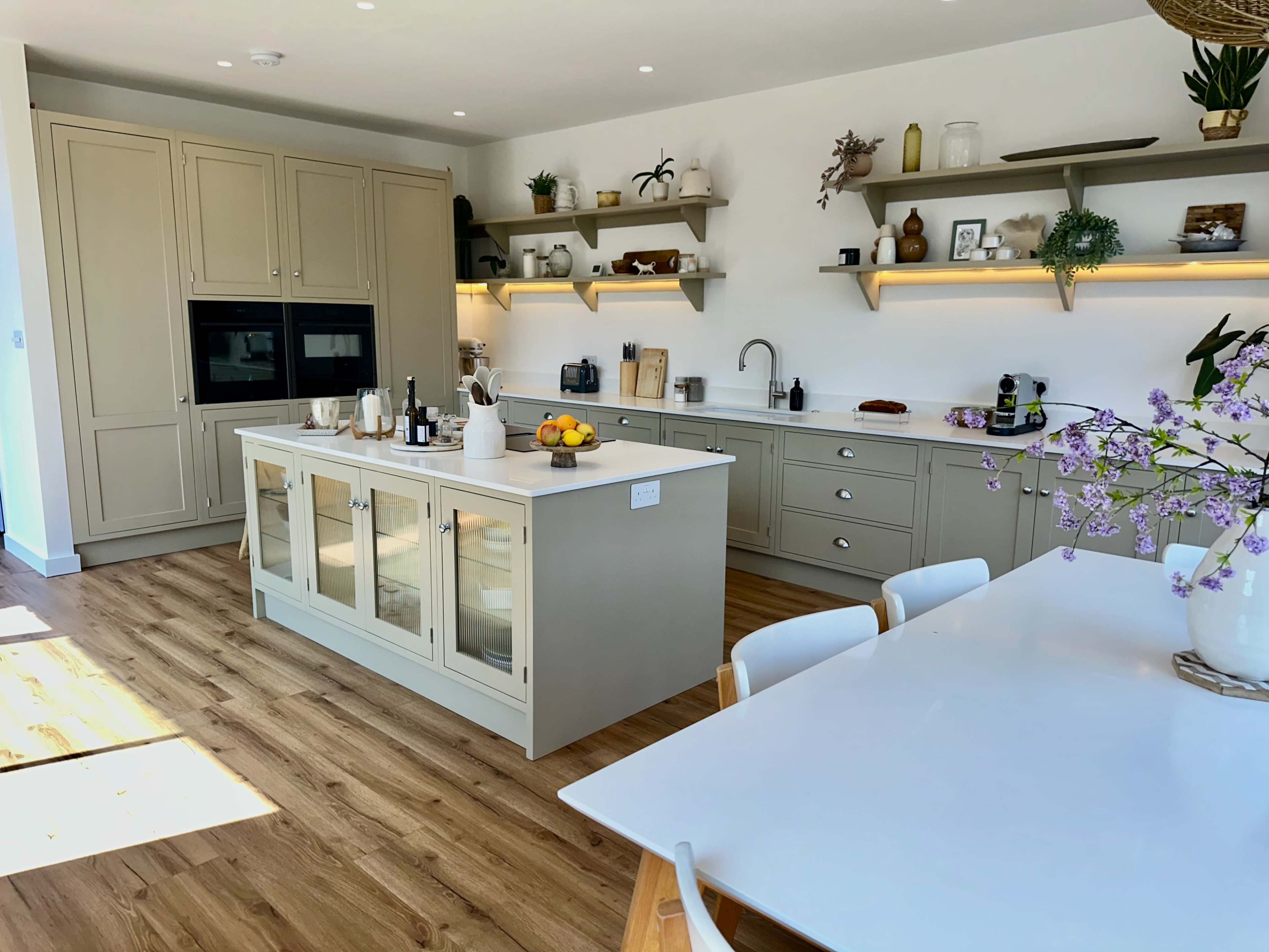 The image shows a modern kitchen with light cabinetry, an island with glass-front cabinets, and open shelving displaying various kitchen items.