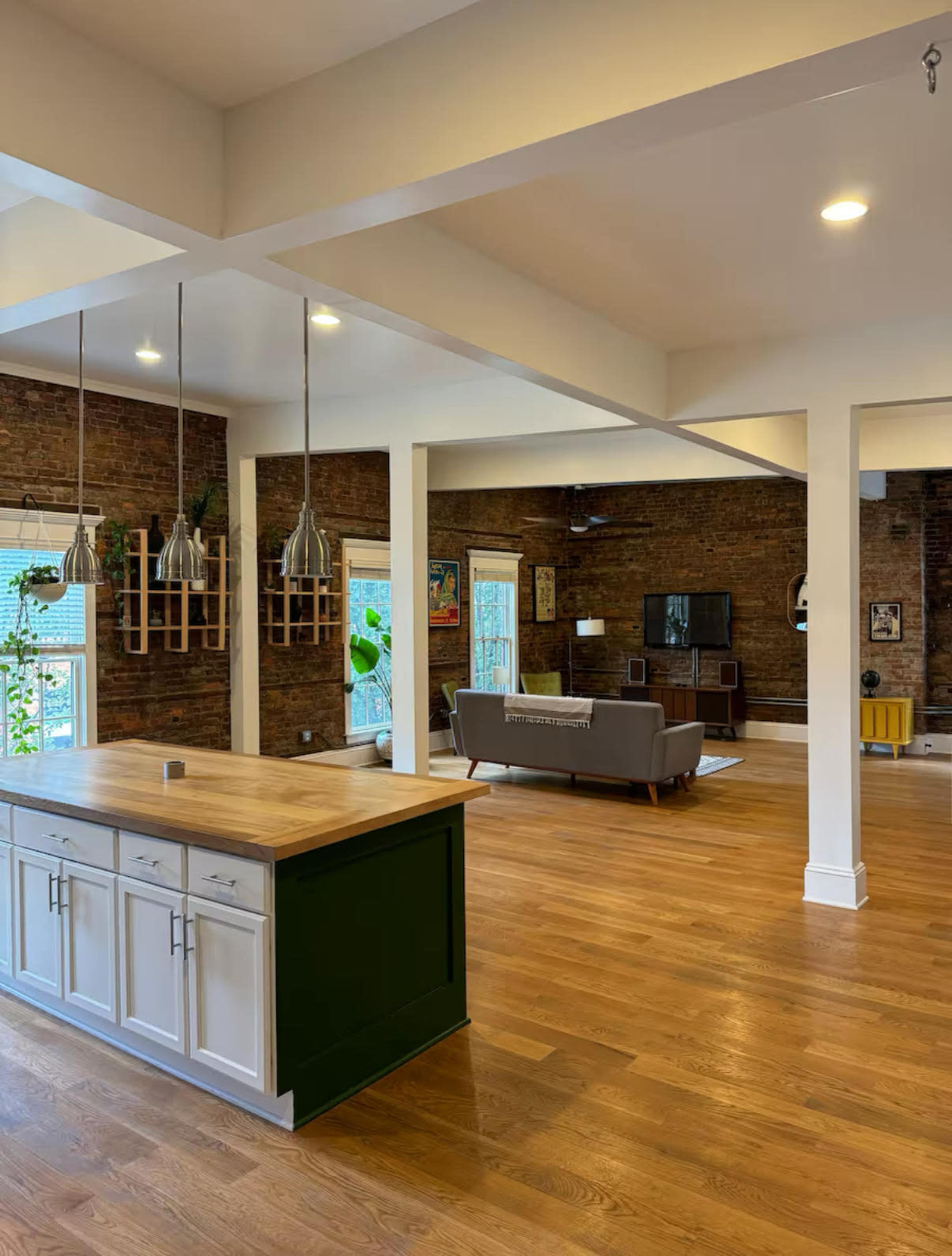 The image shows a spacious living area with brick walls, wooden flooring, and a kitchen island with a green base, leading to a cozy seating area.