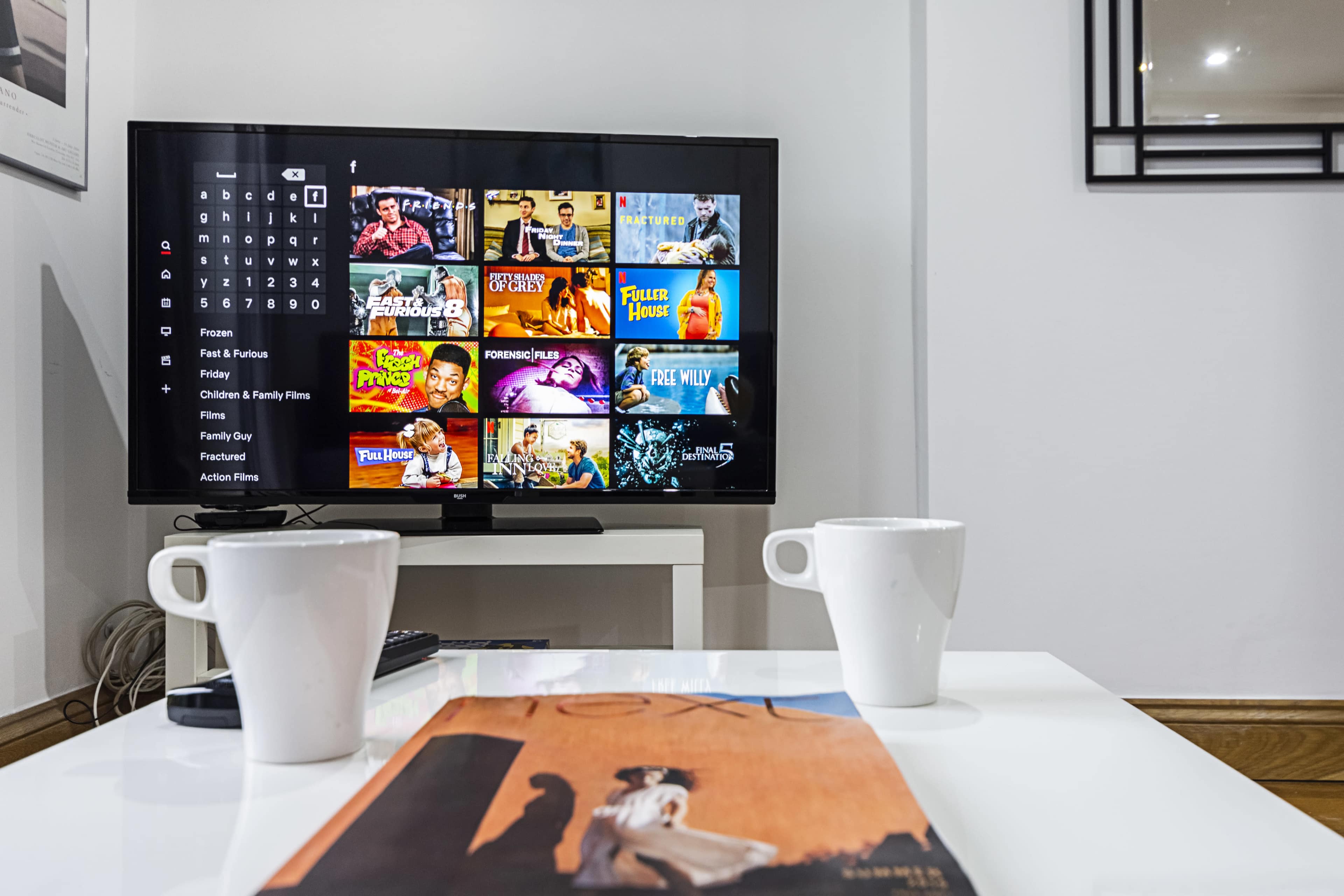 A television displaying a variety of movie and show options is placed on a table with two white mugs and a magazine.