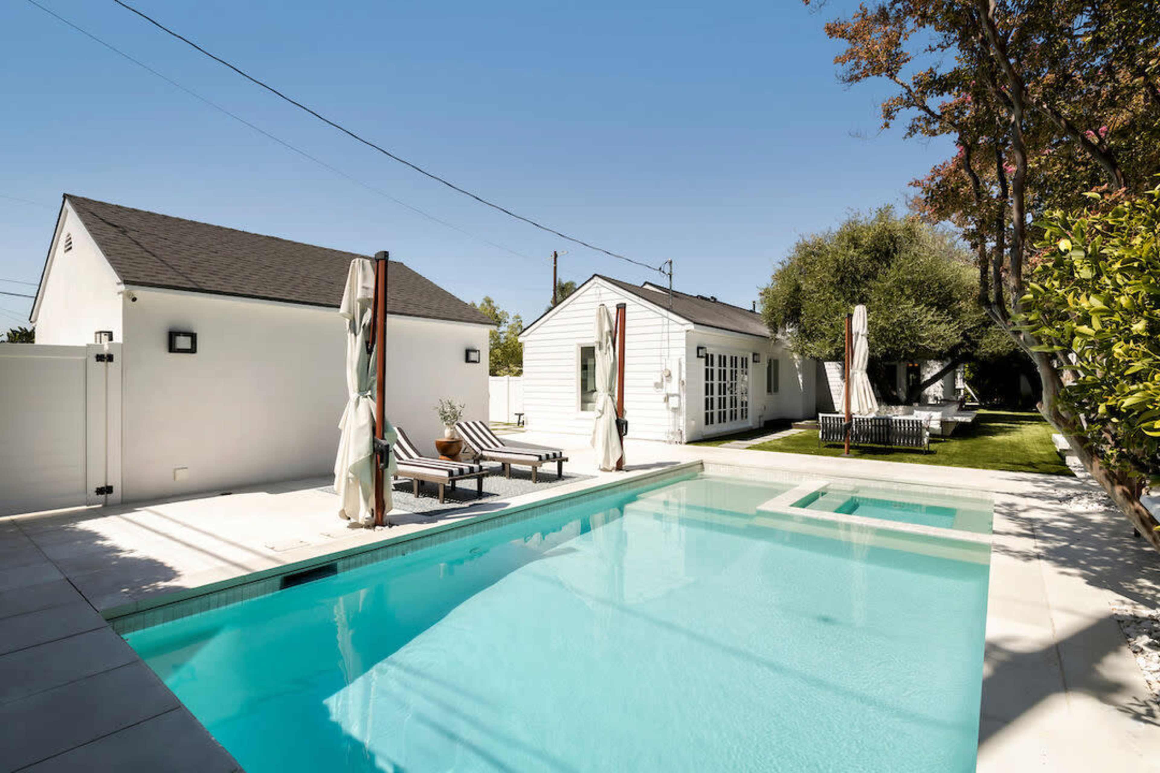 The scene features a modern swimming pool surrounded by lounge chairs and a neatly landscaped backyard with white buildings in the background.