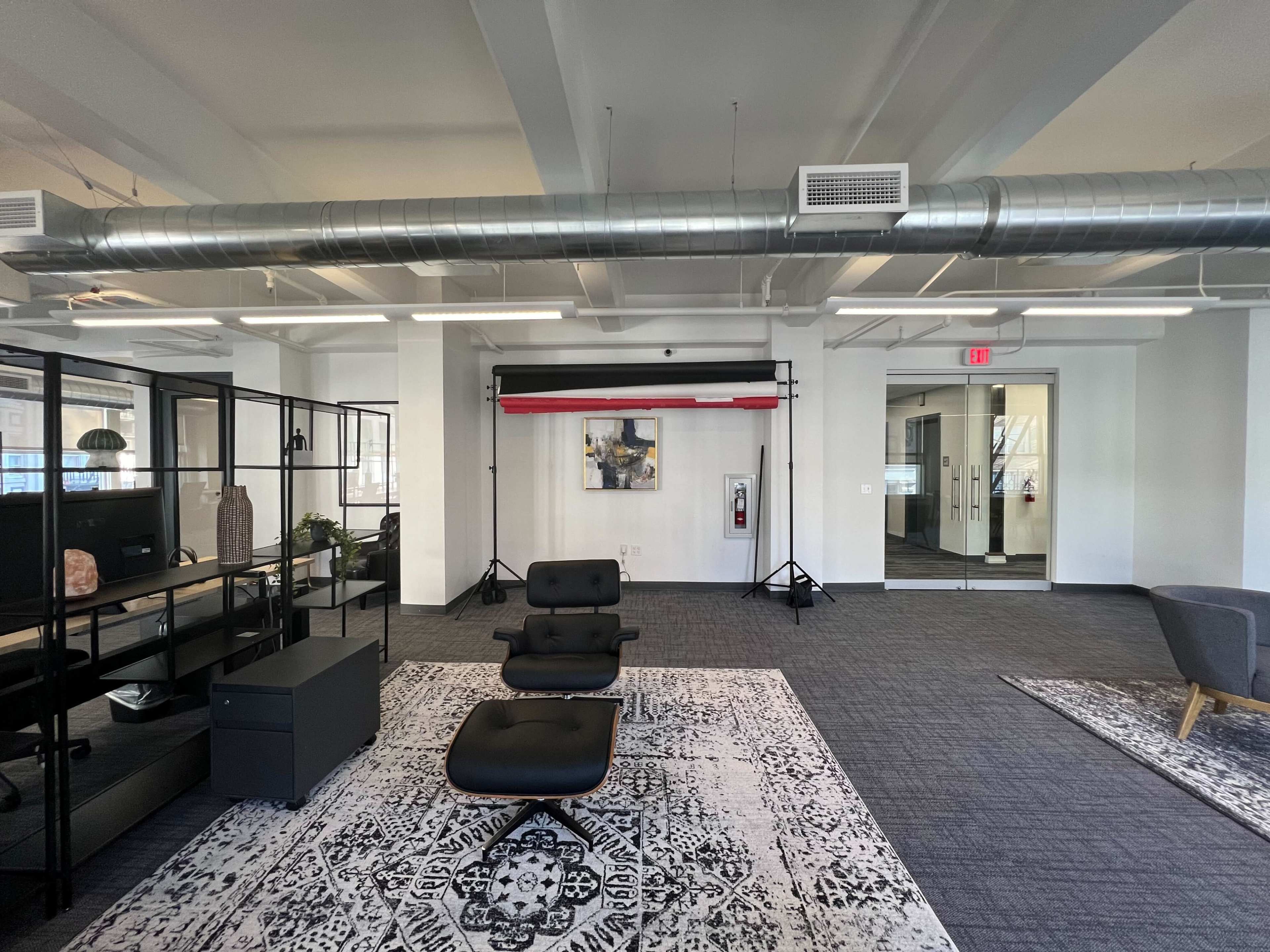 A modern office space with black furniture, a patterned rug, and a backdrop setup near a glass-walled meeting room.