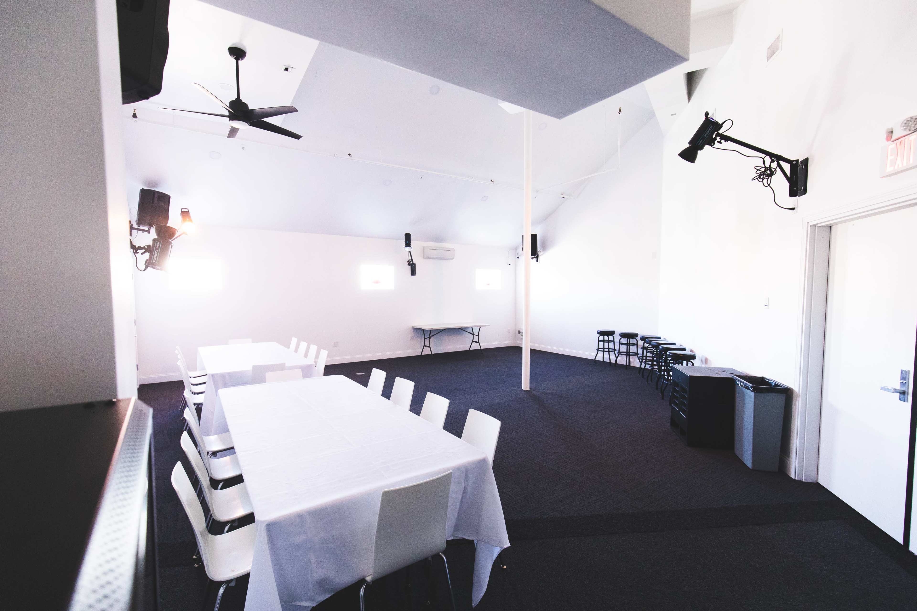 The room features a long table set with white tablecloths, surrounded by chairs, and includes a small stage area with speakers and minimal furnishings.