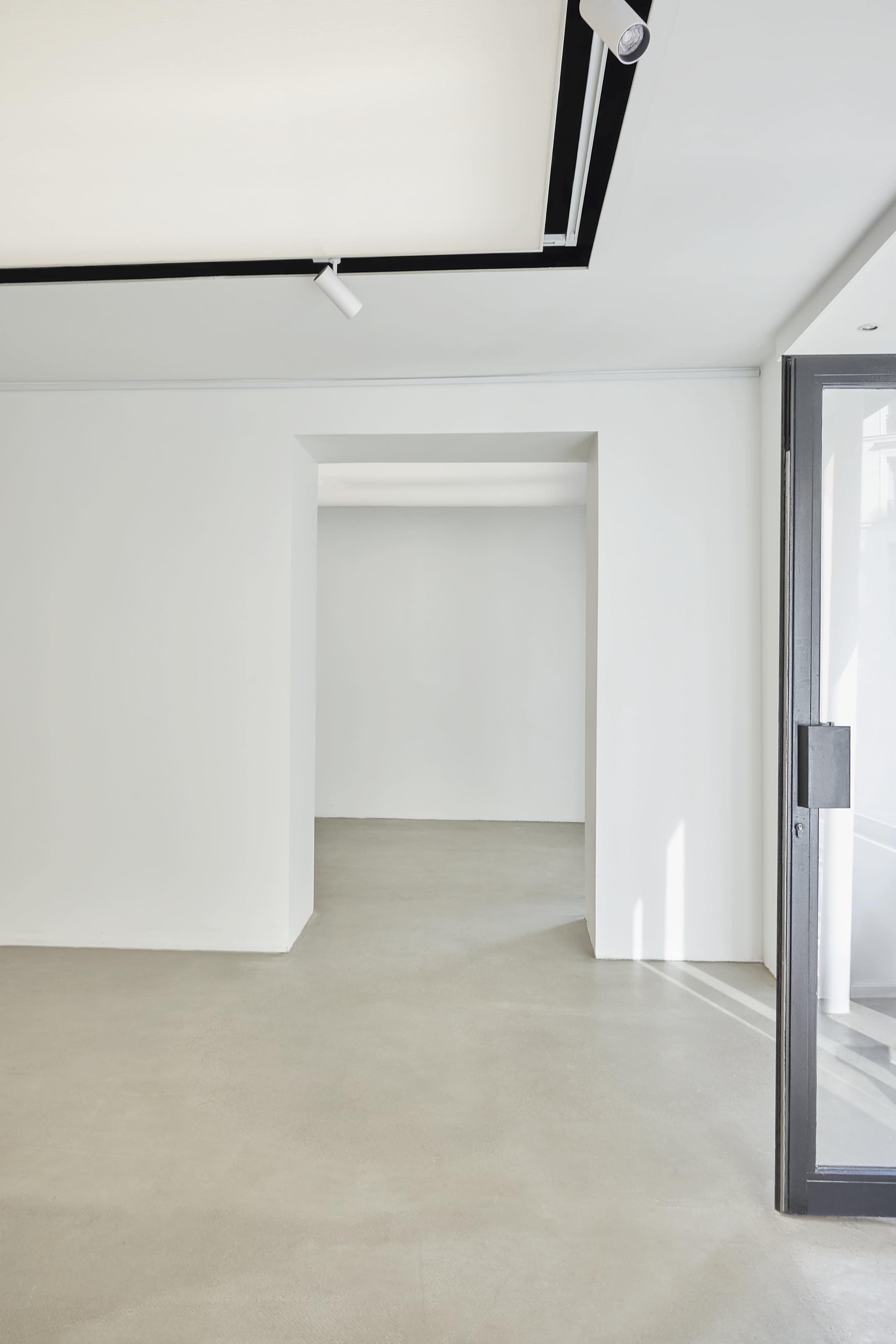 The image shows a minimalistic interior space with white walls, a concrete floor, and an open doorway leading to another room.