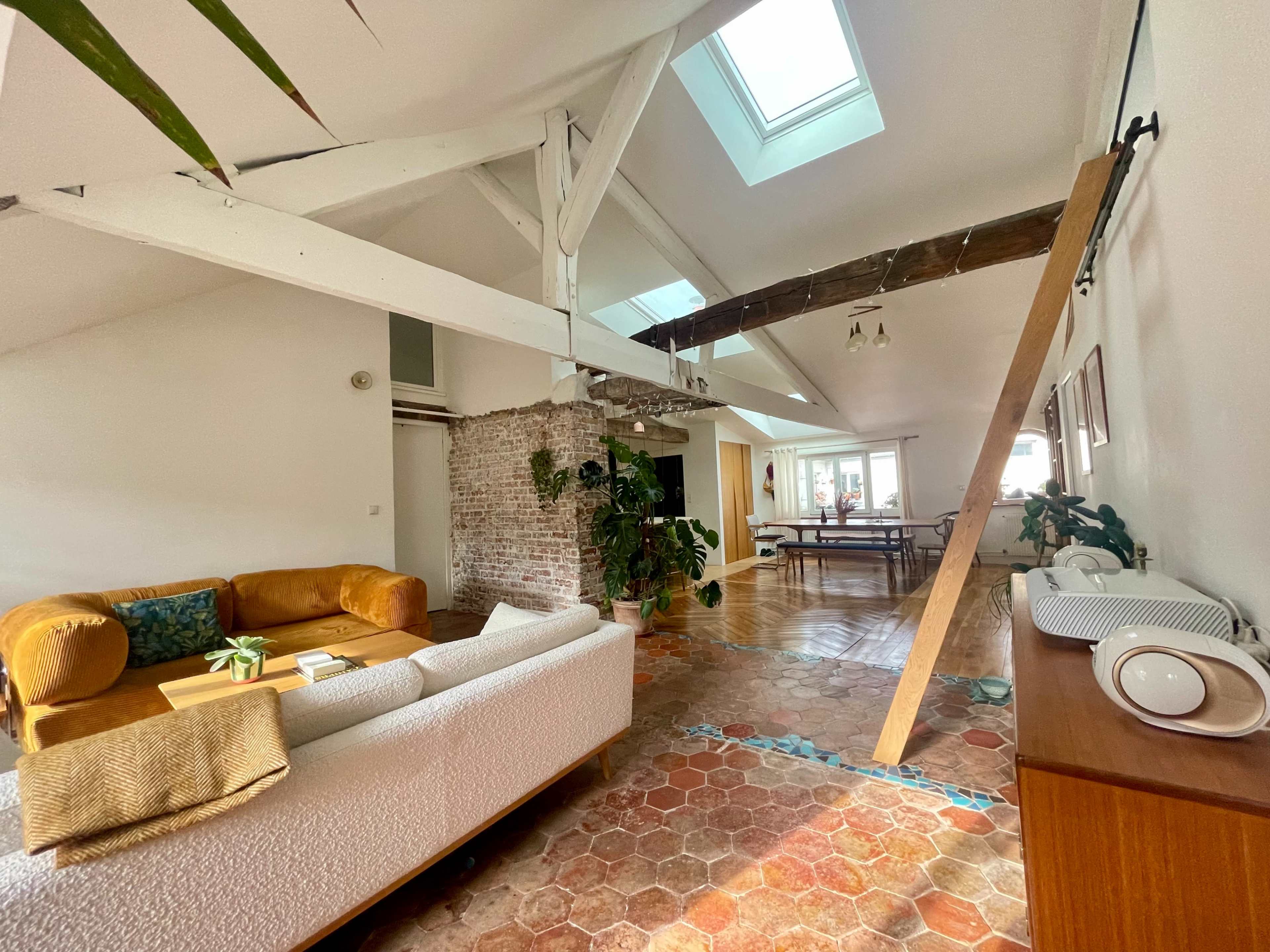 The image shows a spacious, sunlit living area with a couch, a dining table in the background, exposed brick walls, and wooden beams.