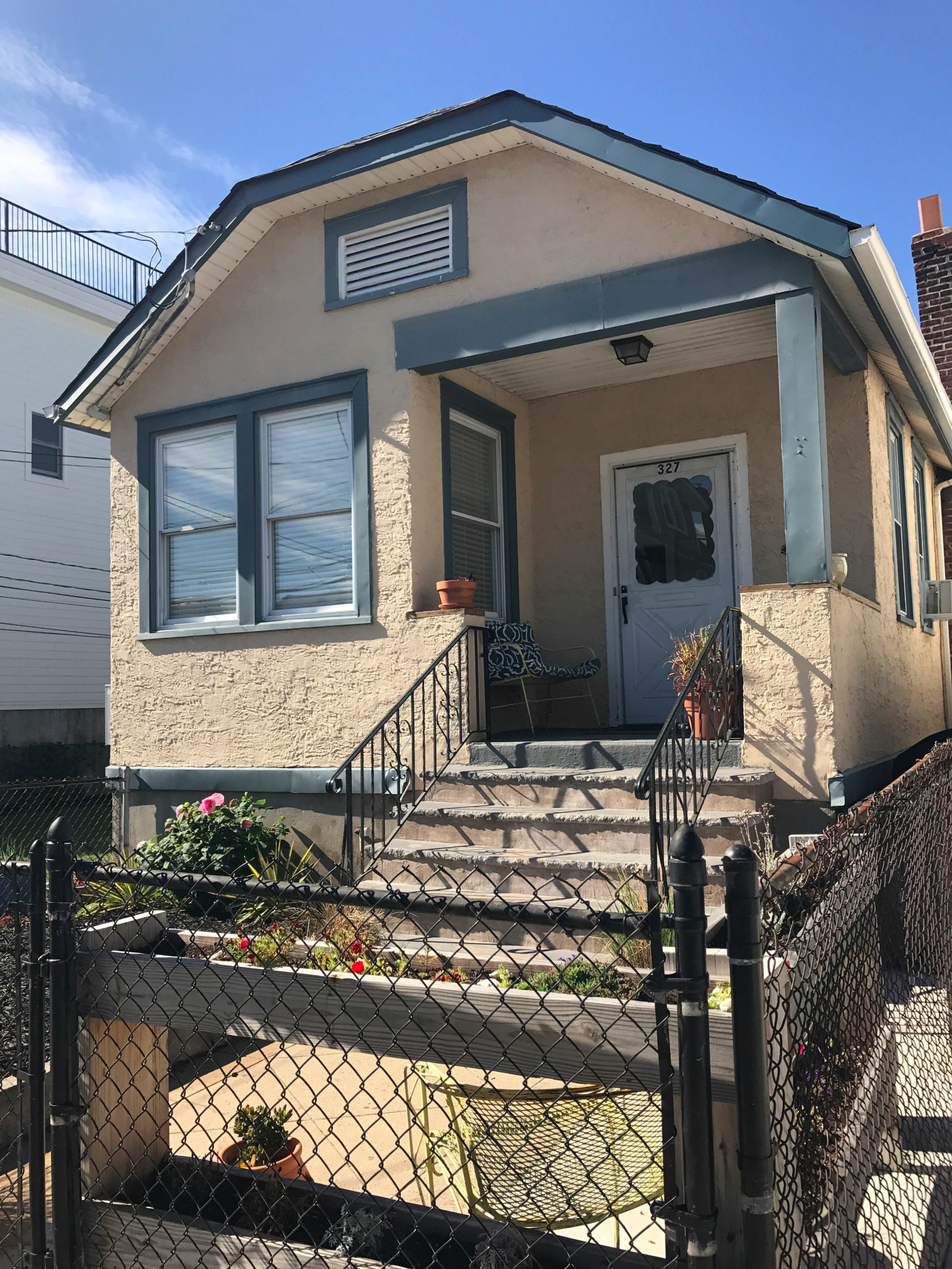 The image shows a small, single-story house with a front porch, surrounded by a fence and featuring a garden area.