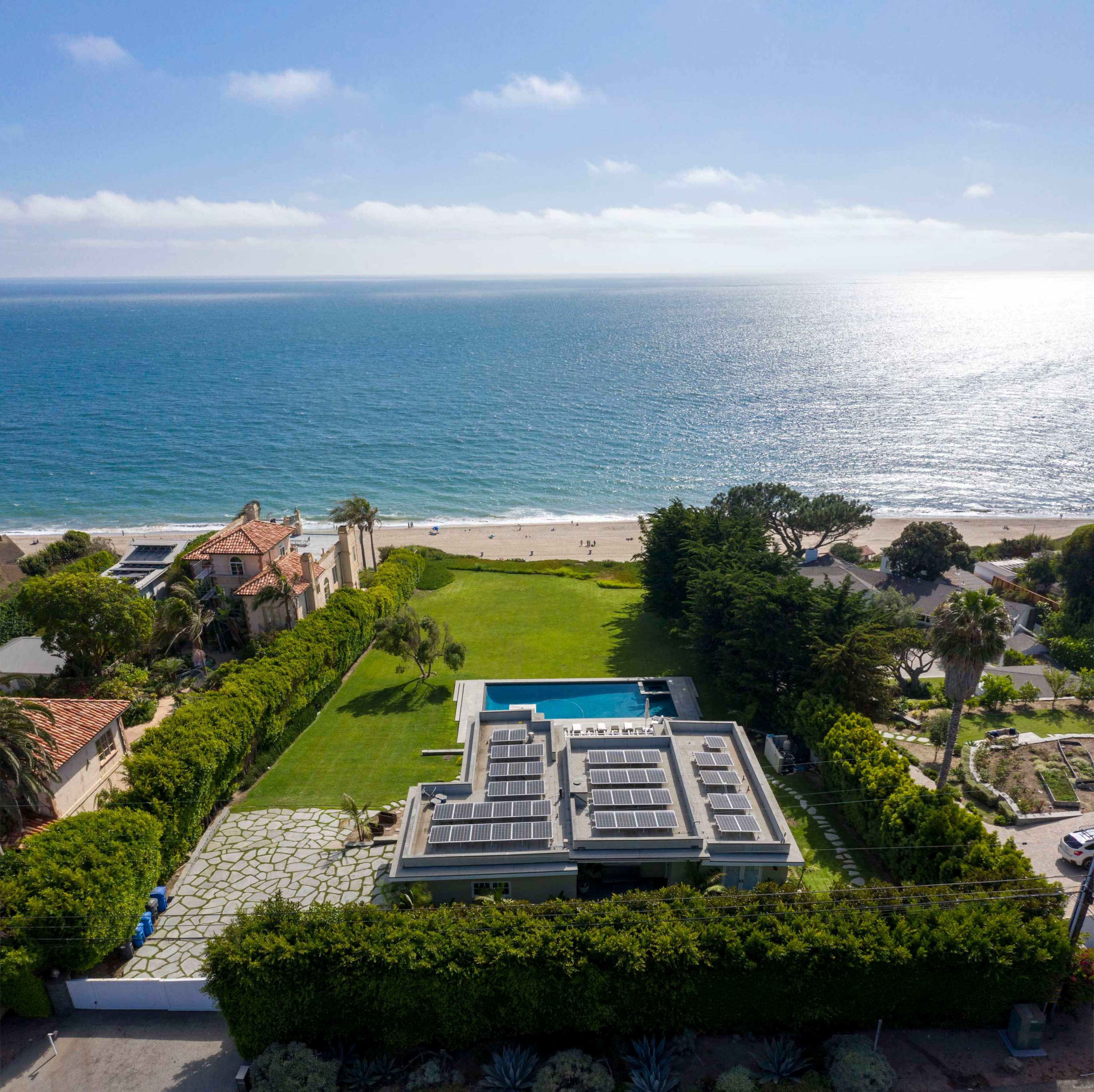 An aerial view shows a beachfront house with a green lawn, a swimming pool, and solar panels on the roof, adjacent to a sandy beach and the ocean in the background.