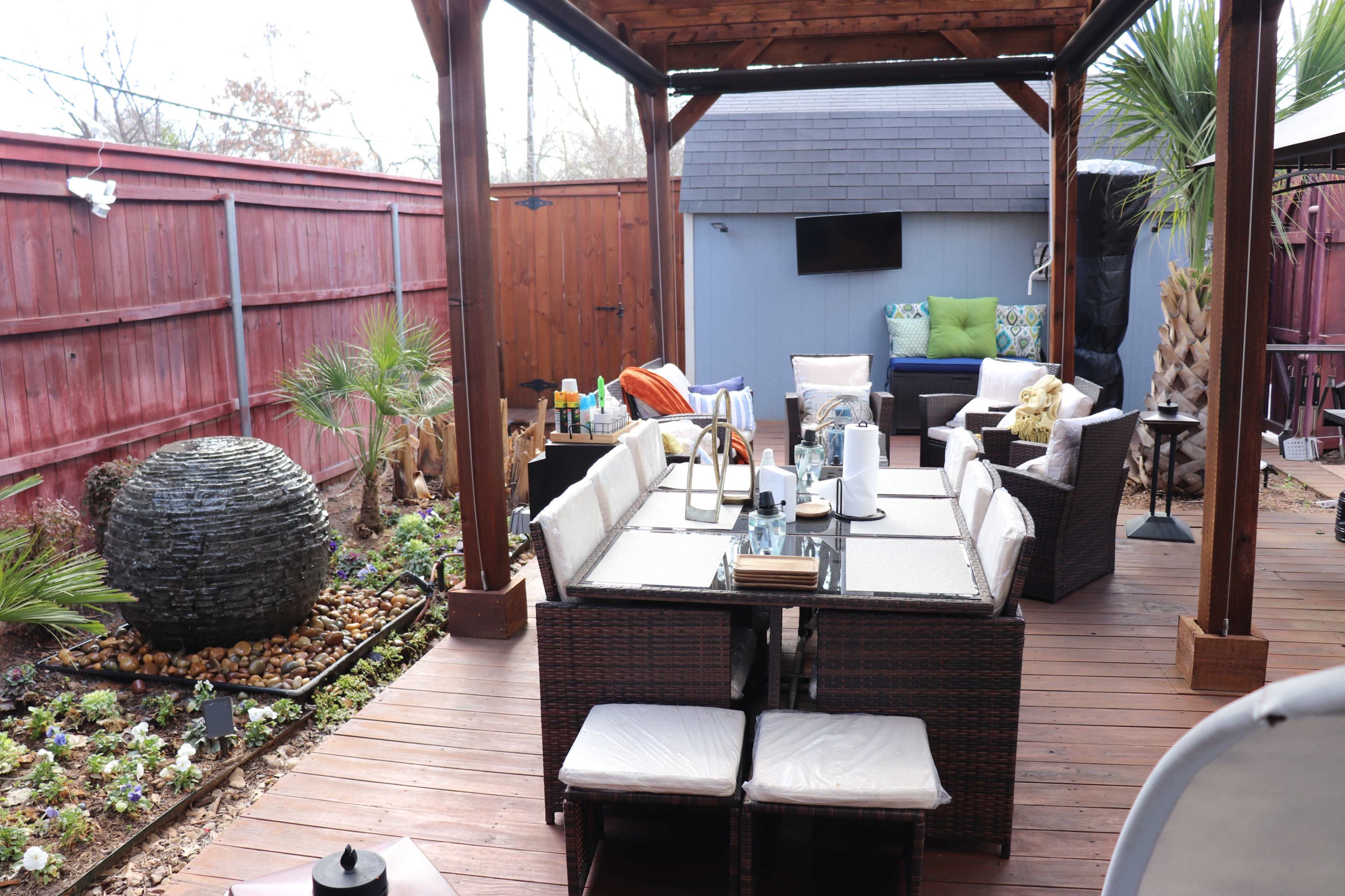 The image shows a wooden deck patio with a seating area of wicker furniture, a large decorative sphere, and potted plants.