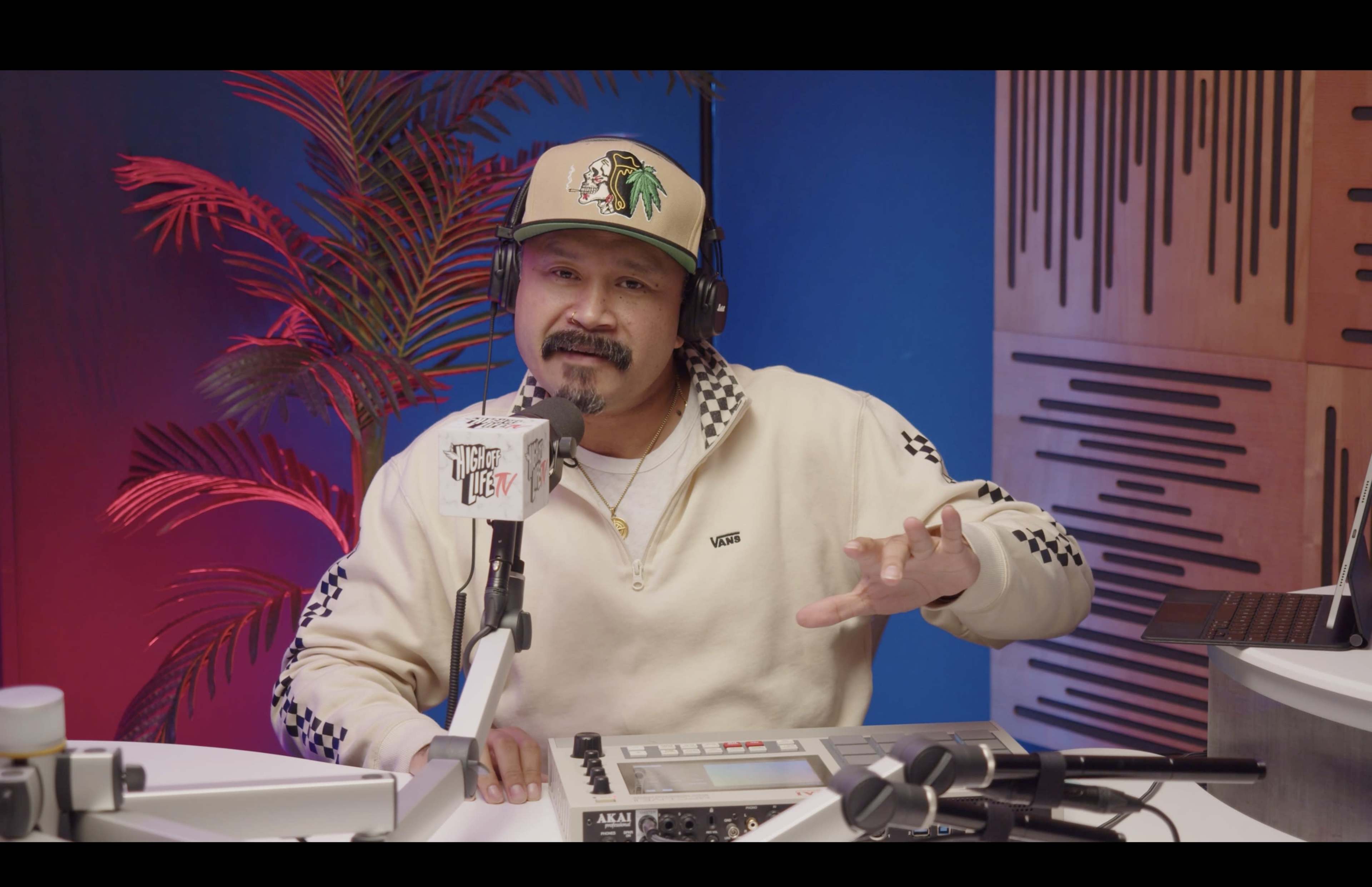 A man wearing a beige cap and a white sweatshirt is speaking into a microphone in a colorful studio setting.