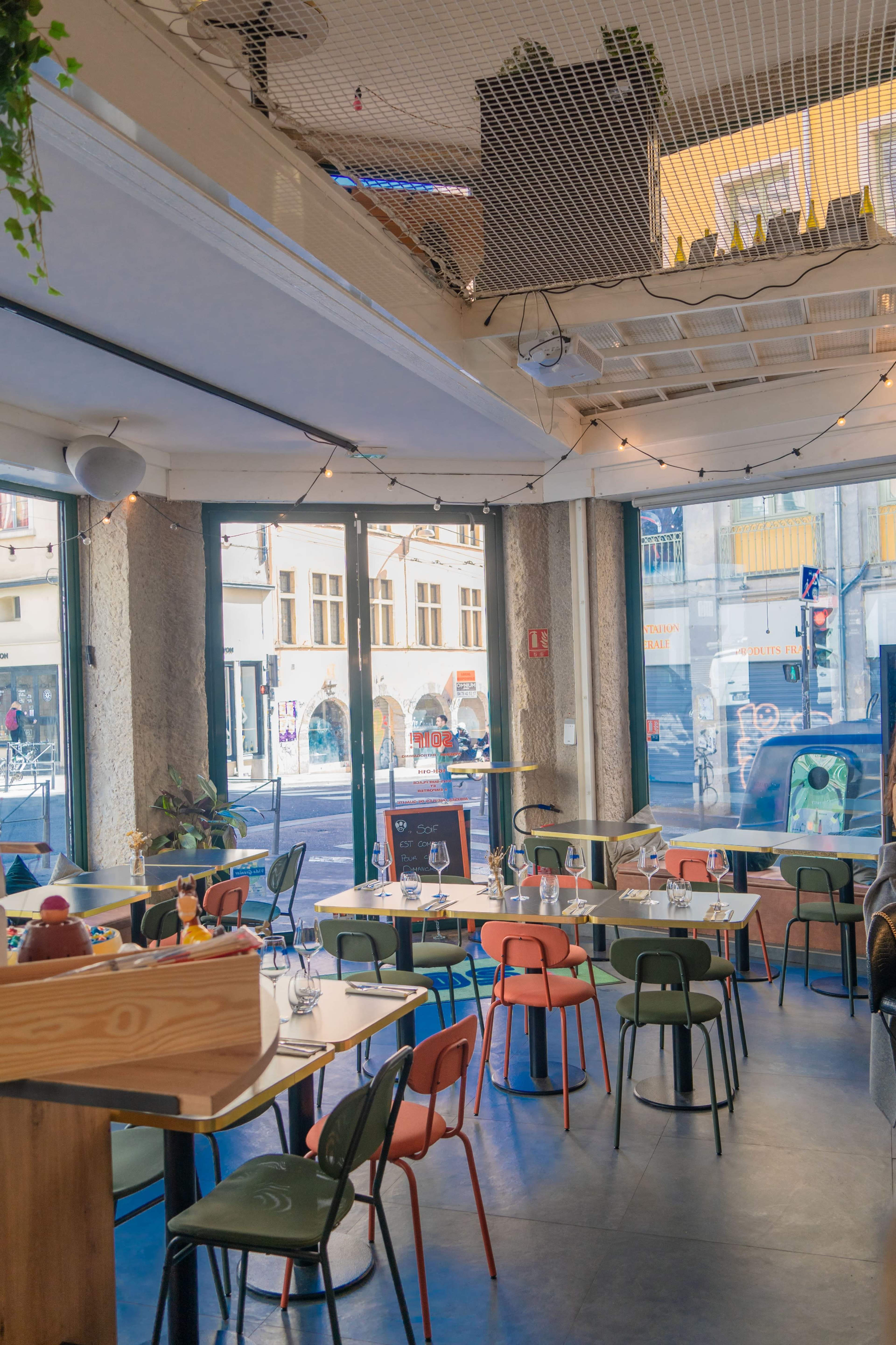 The image shows a modern café interior with colorful chairs and tables, large windows allowing natural light, and a view of the street outside.