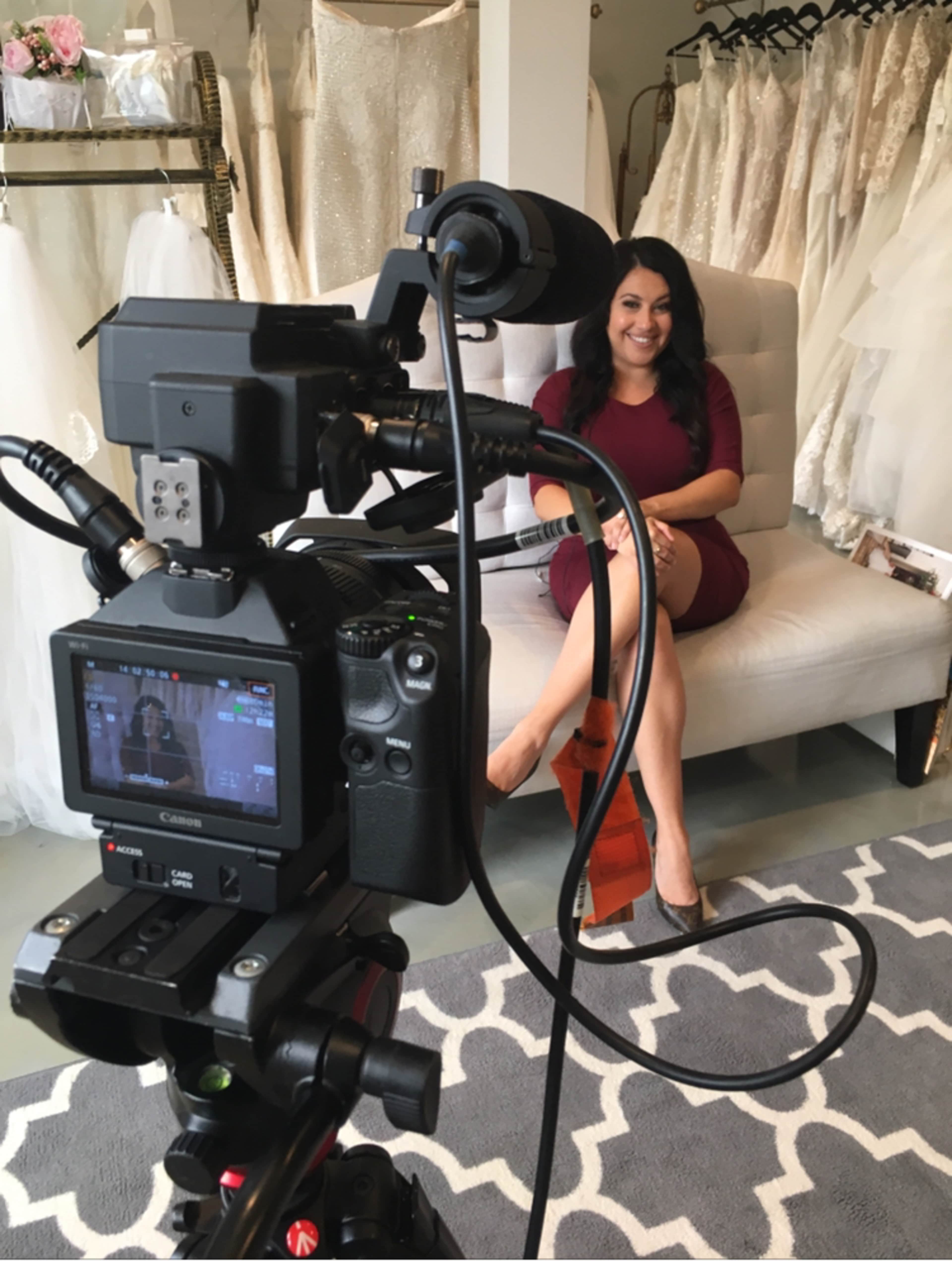 A camera is set up in a bridal shop, focusing on a woman sitting on a couch among wedding dresses on display.