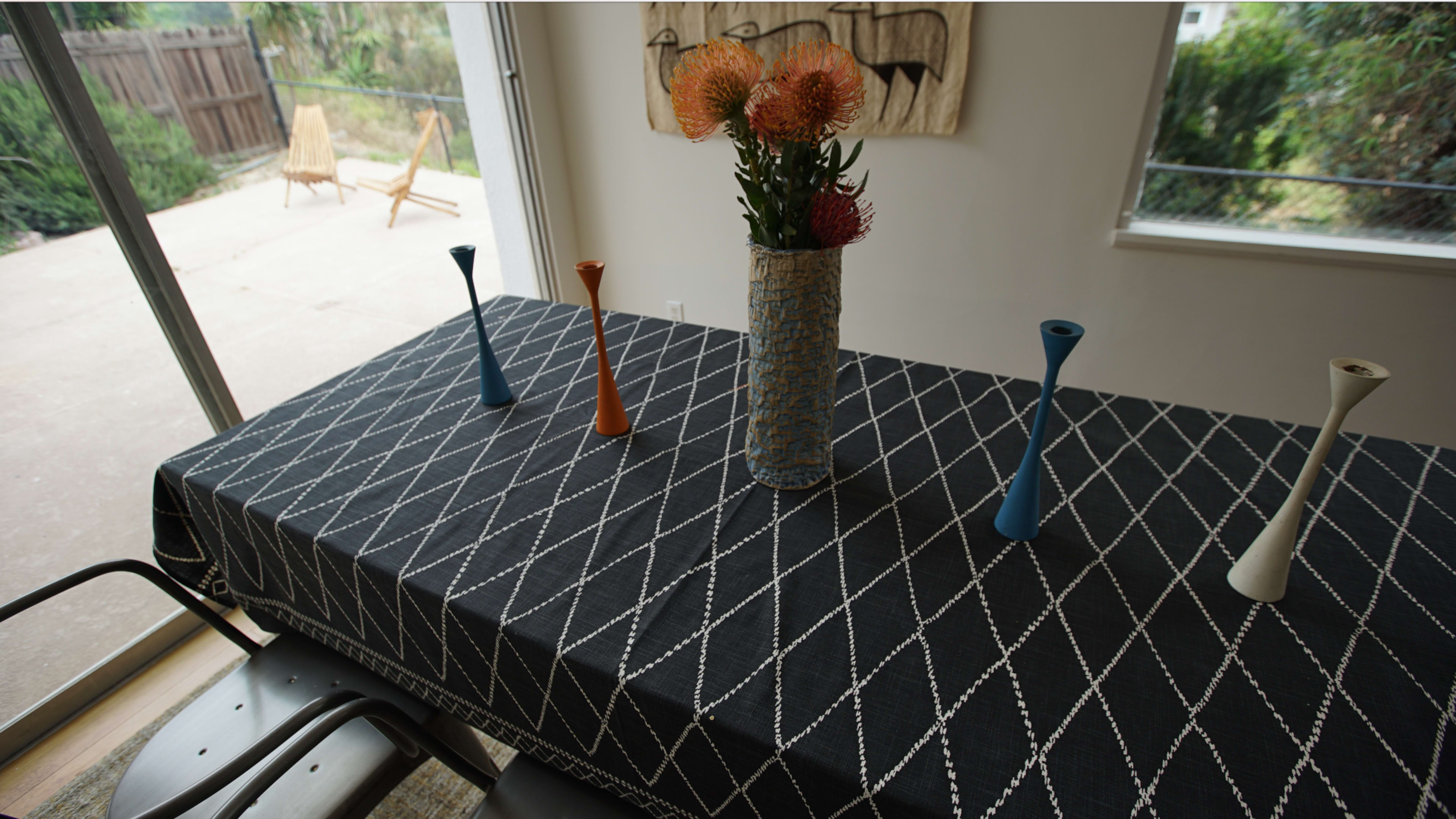 A patterned black tablecloth covers a dining table with a vase of flowers and several colorful vases arranged on top.