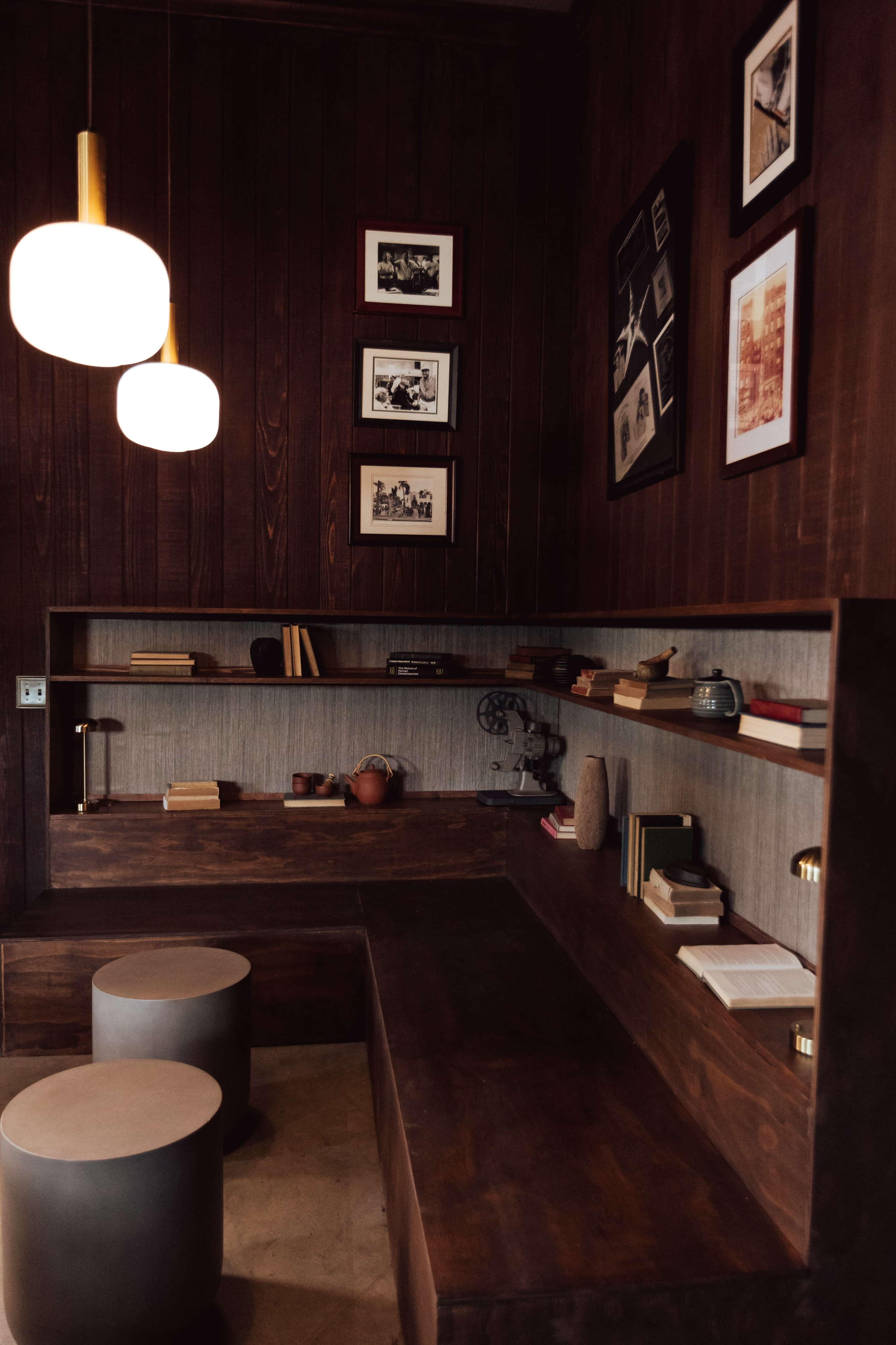 A cozy interior featuring wooden shelves with books and decorative items, two round stools, and framed photographs on the wall, illuminated by hanging pendant lights.