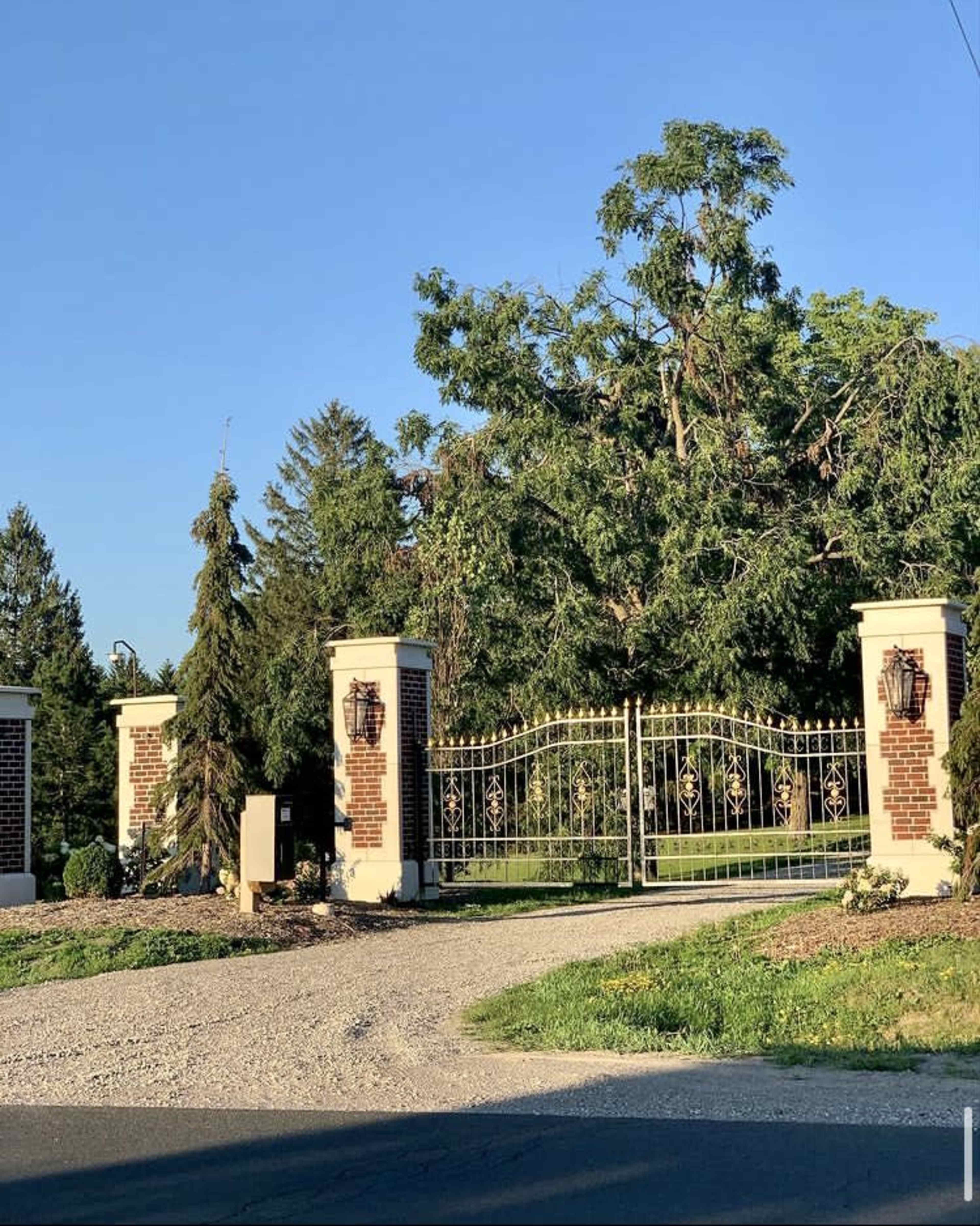 A gated entrance with brick pillars and a decorative metal gate, surrounded by trees and a gravel driveway.
