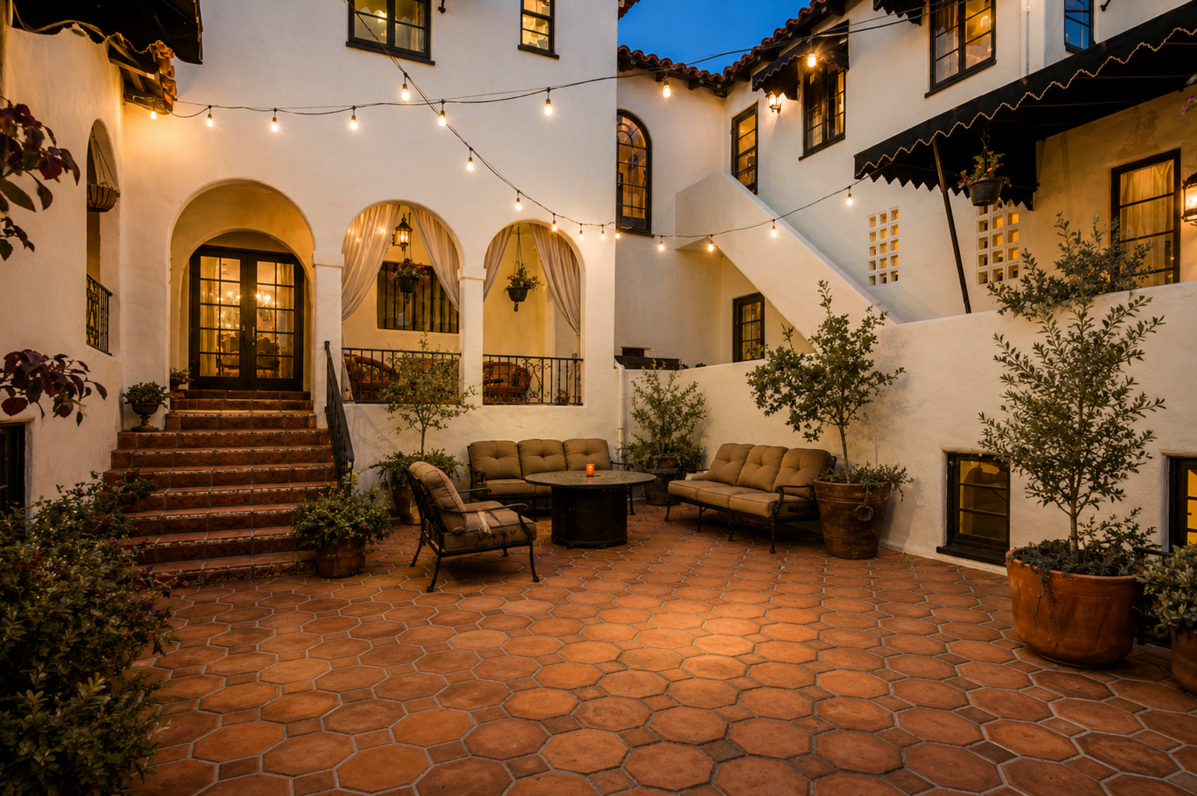 The image shows a courtyard with terracotta tiles, surrounded by white stucco walls, featuring string lights, outdoor seating, and potted plants.