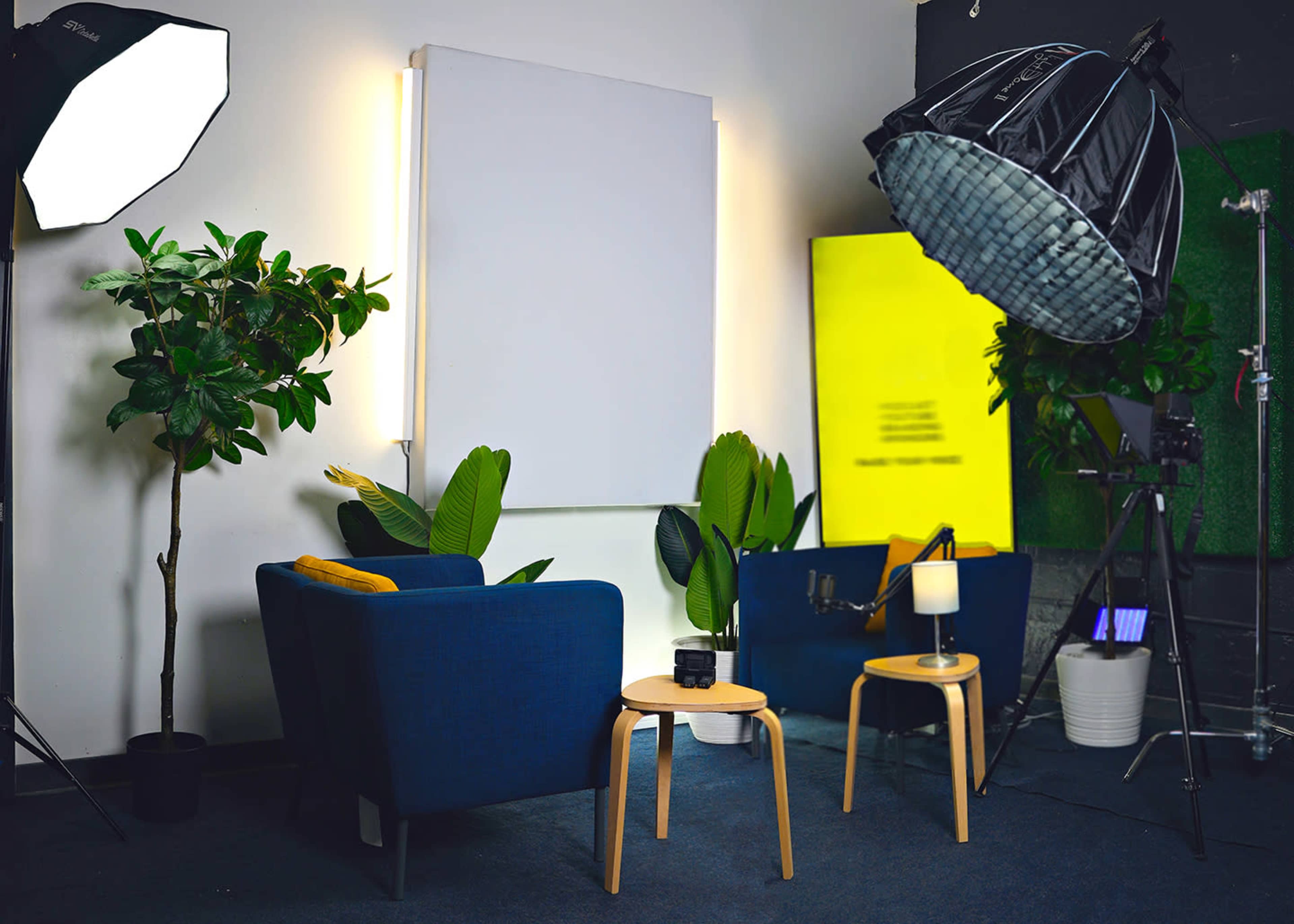 The image depicts a modern interview setup featuring two blue chairs, a small table, and various lighting equipment against a backdrop of plants and a blank white canvas.