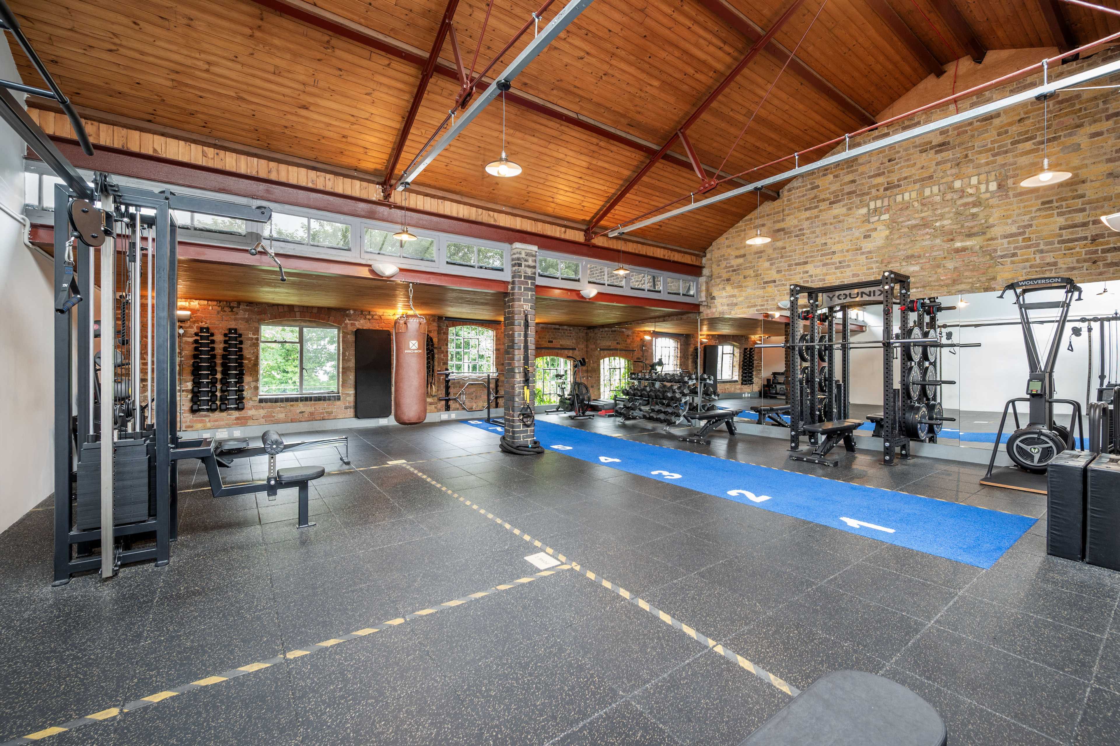 The image shows a spacious gym with various exercise equipment, including weight machines, free weights, and a cable station, all set against a backdrop of brick walls and large windows.