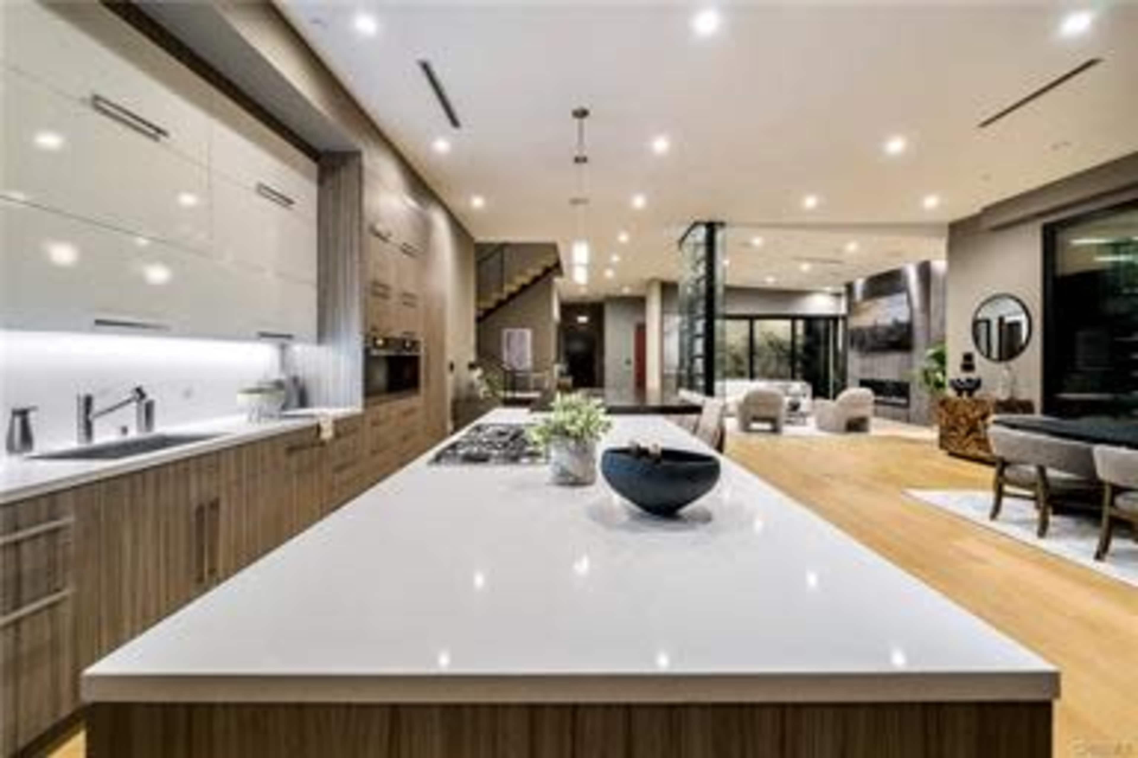 A modern kitchen features a large island with a cooktop, surrounded by sleek cabinetry and an open layout leading to a living area.