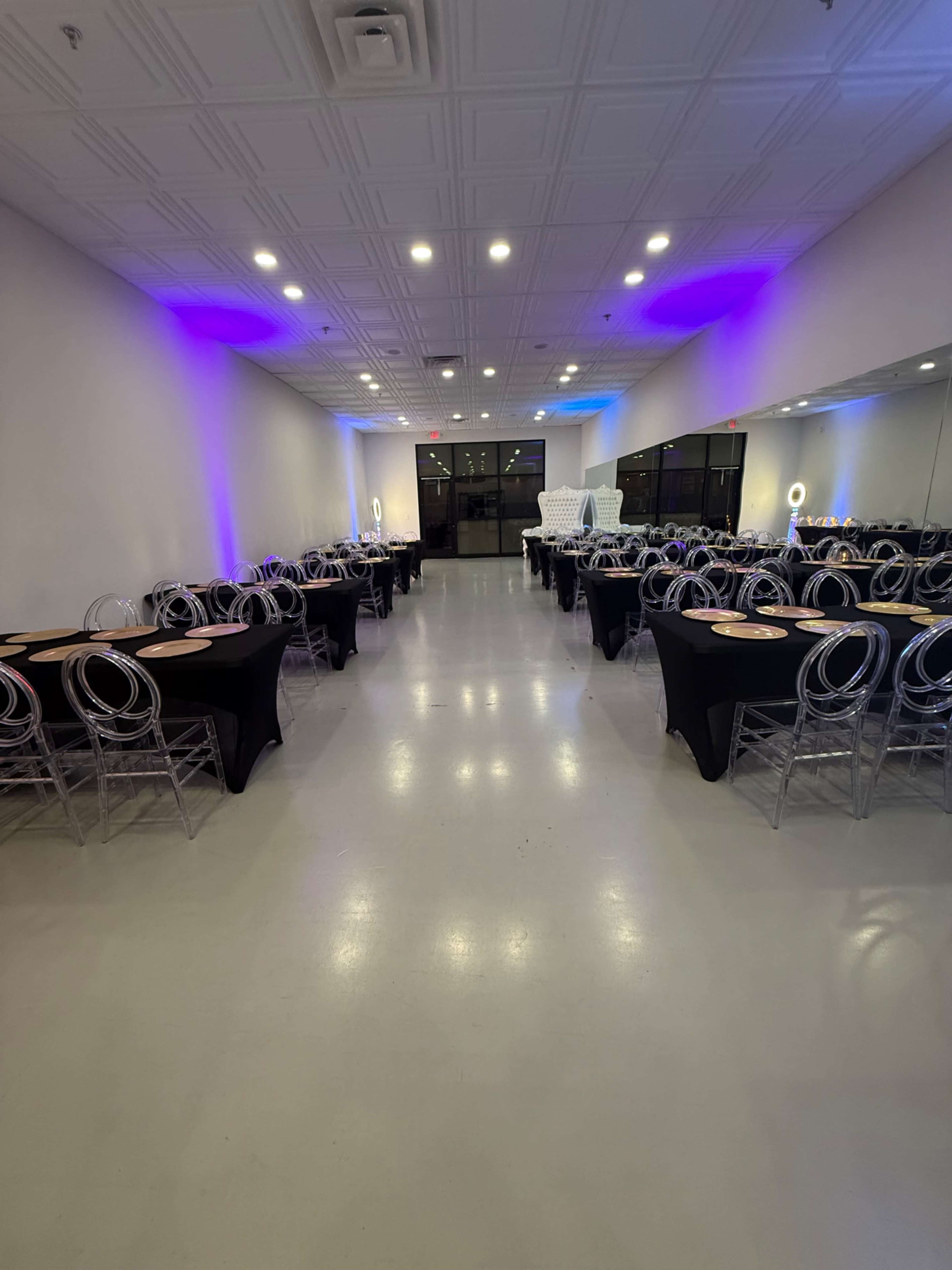A spacious event room features rows of black-covered tables with transparent chairs and purple lights illuminating the walls.