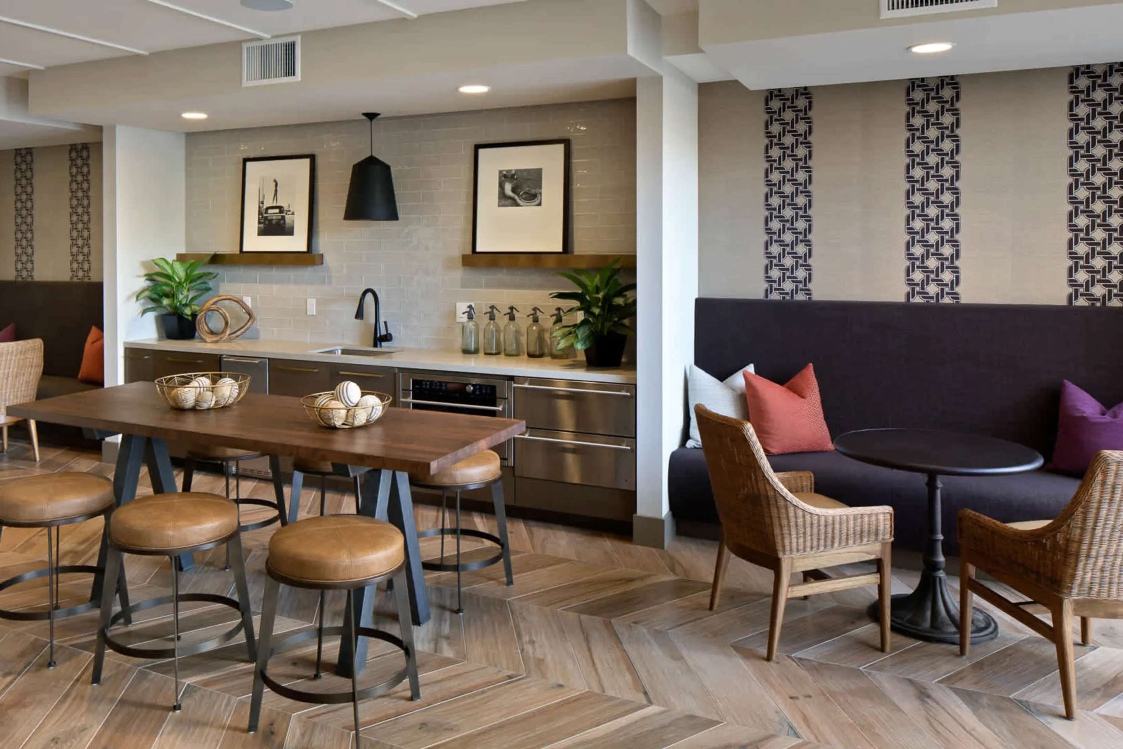 A modern kitchen and dining area featuring sleek cabinetry, a large wooden table with stools, and a cozy seating nook against a patterned wall.