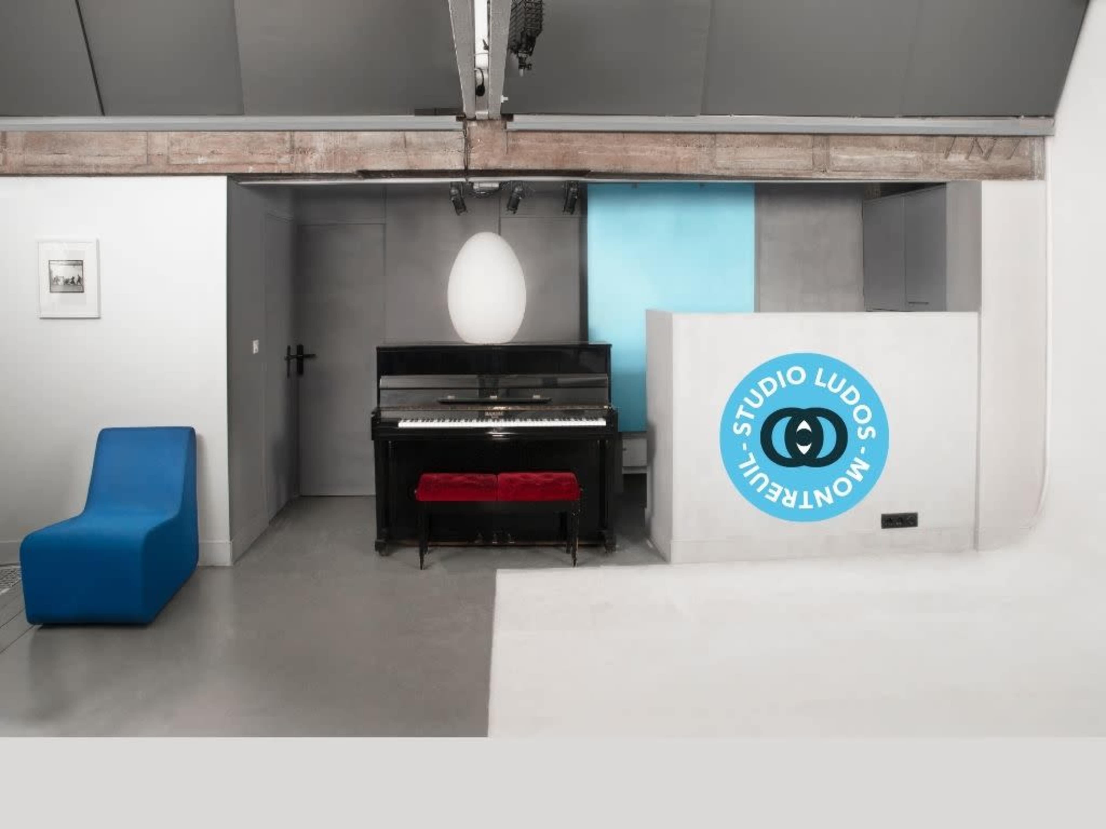 The image depicts a modern studio space featuring a black piano, a red bench, a blue accent wall, and a reception area with a circular logo.