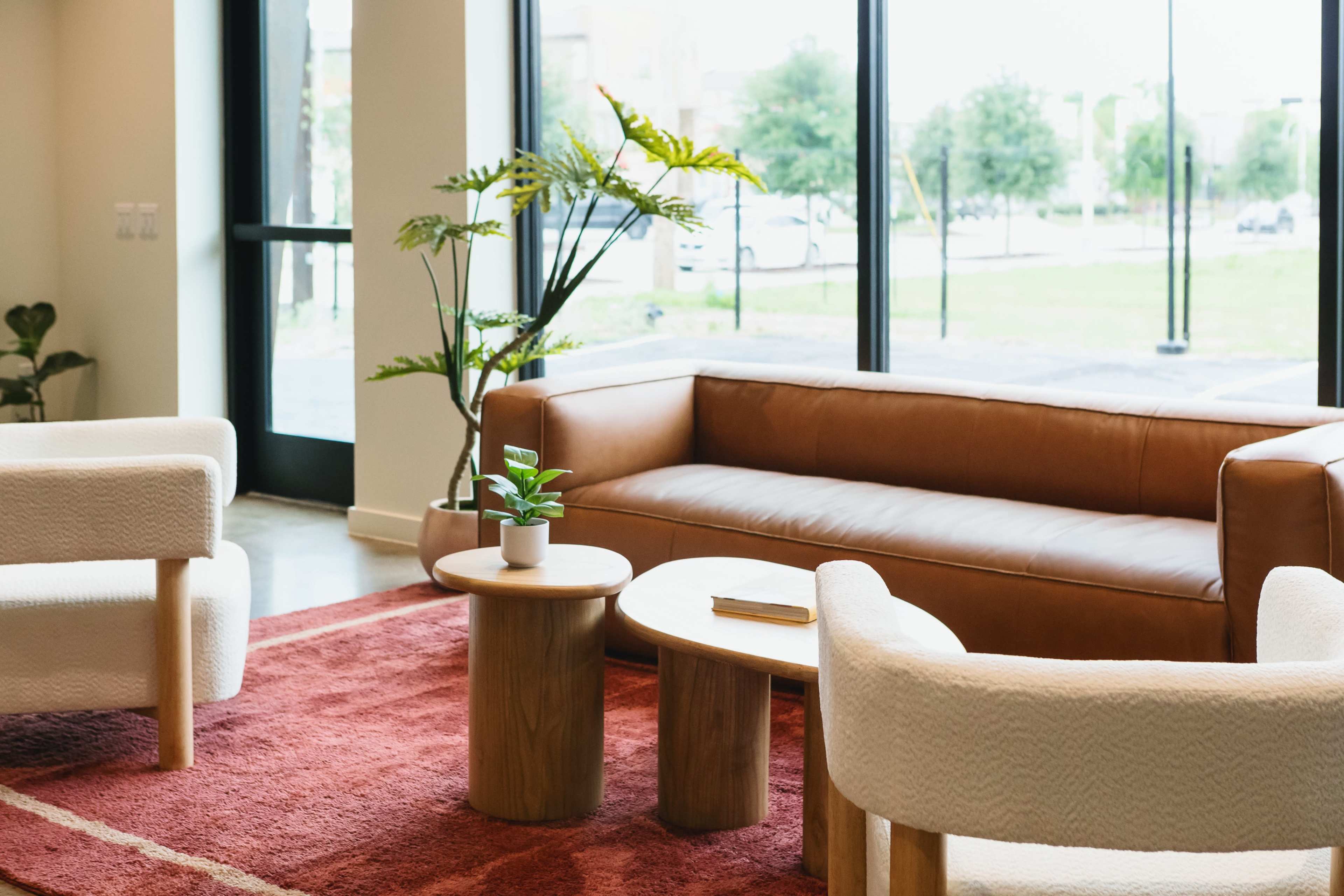 A modern living area features a brown leather sofa, two white upholstered chairs, a small round wooden table, and a potted plant, with large windows allowing natural light to fill the space.
