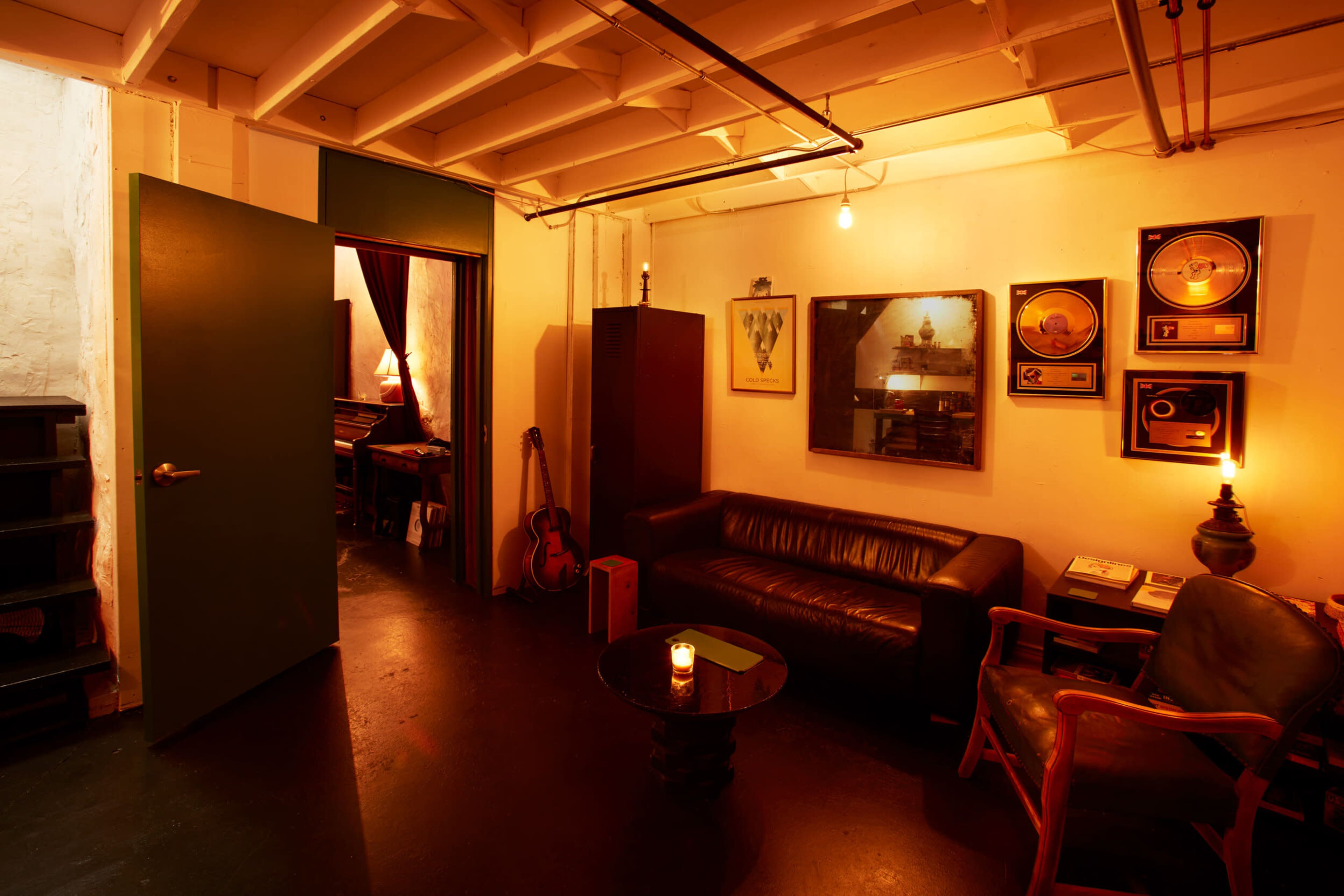 A cozy room with a sofa, a table with a candle, a guitar, and framed records on the wall, with an open door leading to a darker space.