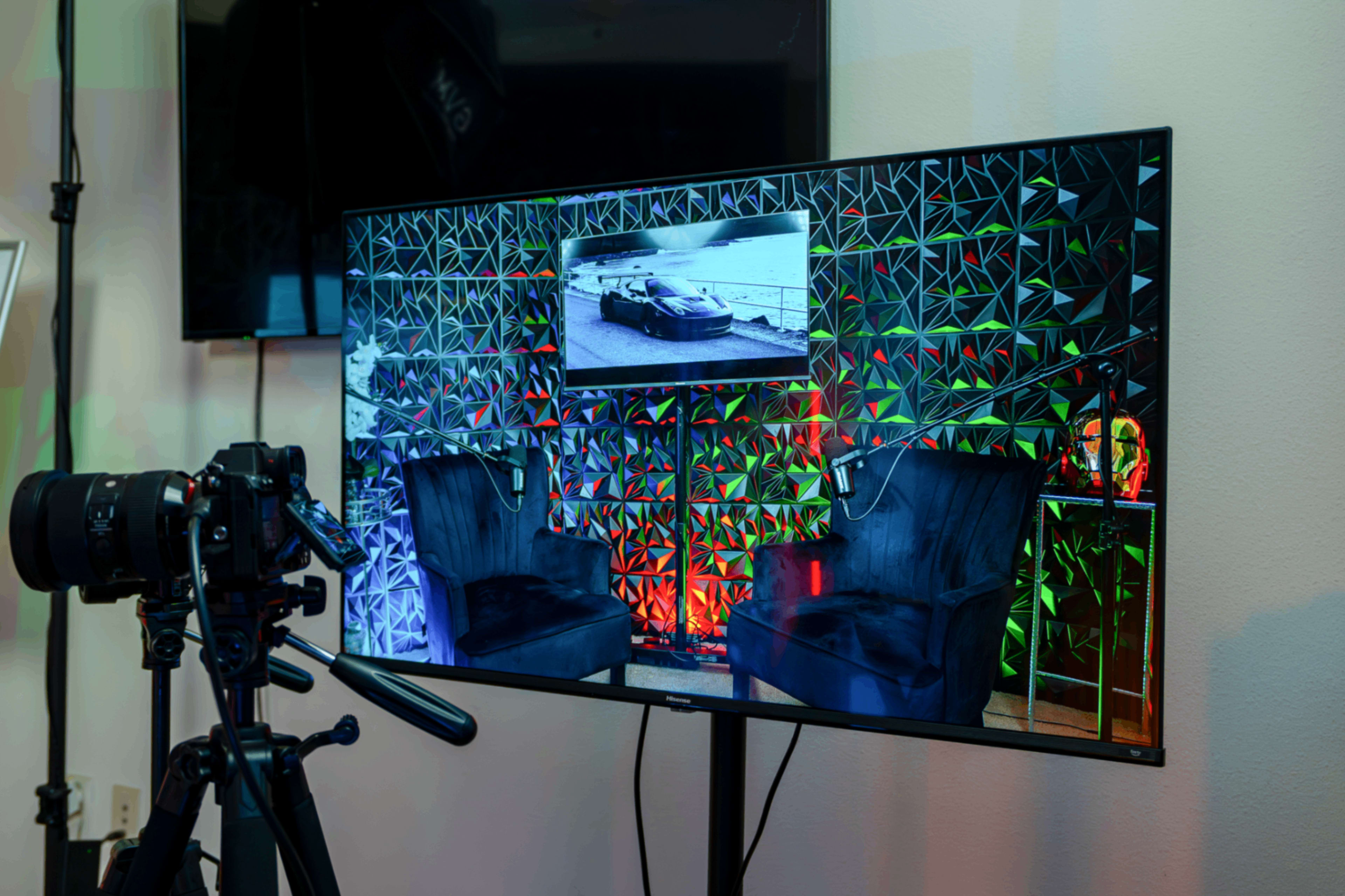 The image shows a camera on a tripod facing a large screen displaying a car video, with two black chairs set in front of a textured, colorful backdrop.