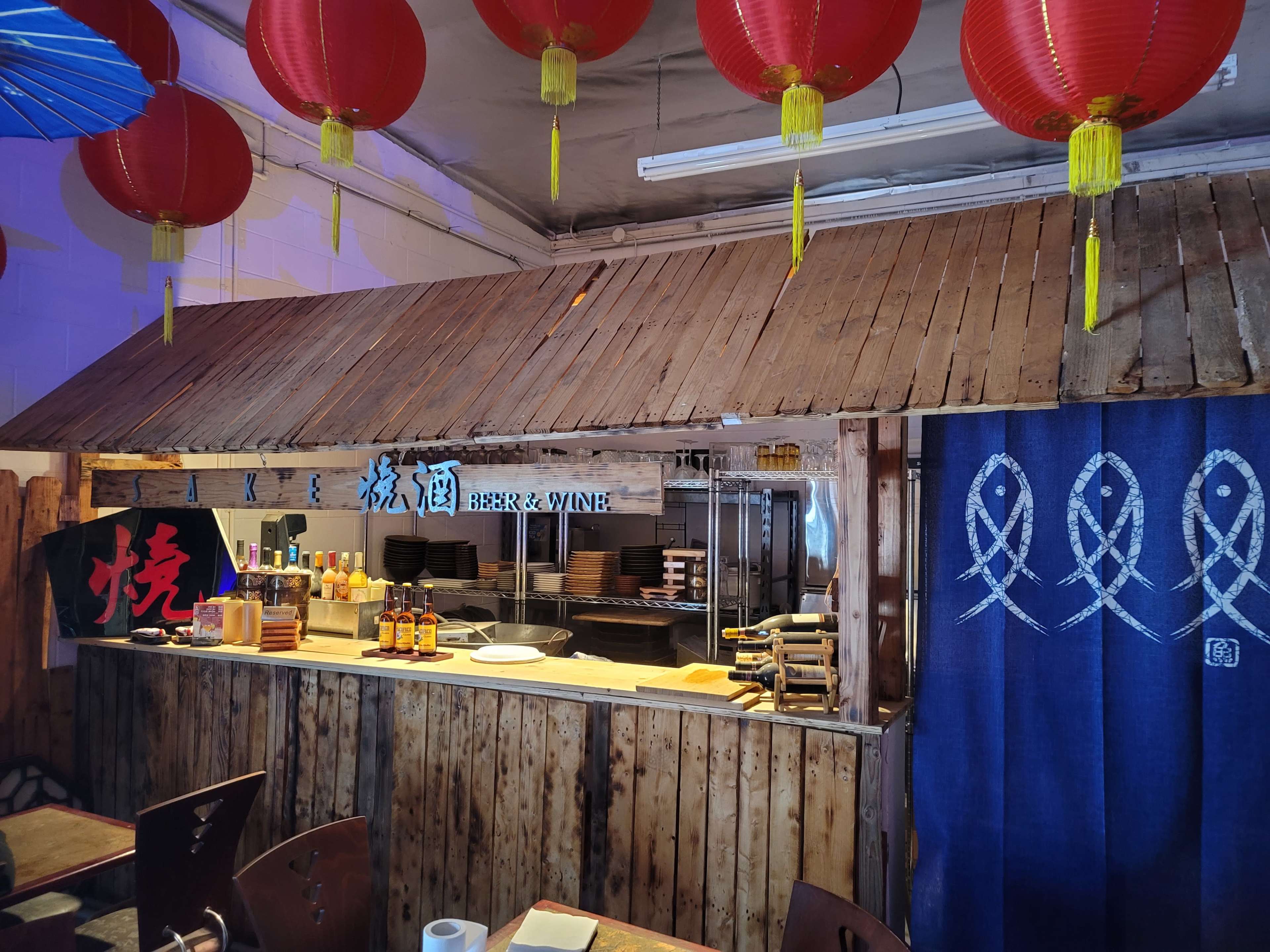 The interior of a restaurant featuring a wooden counter, traditional decorations including red lanterns, and a blue fabric curtain with decorative fish patterns.