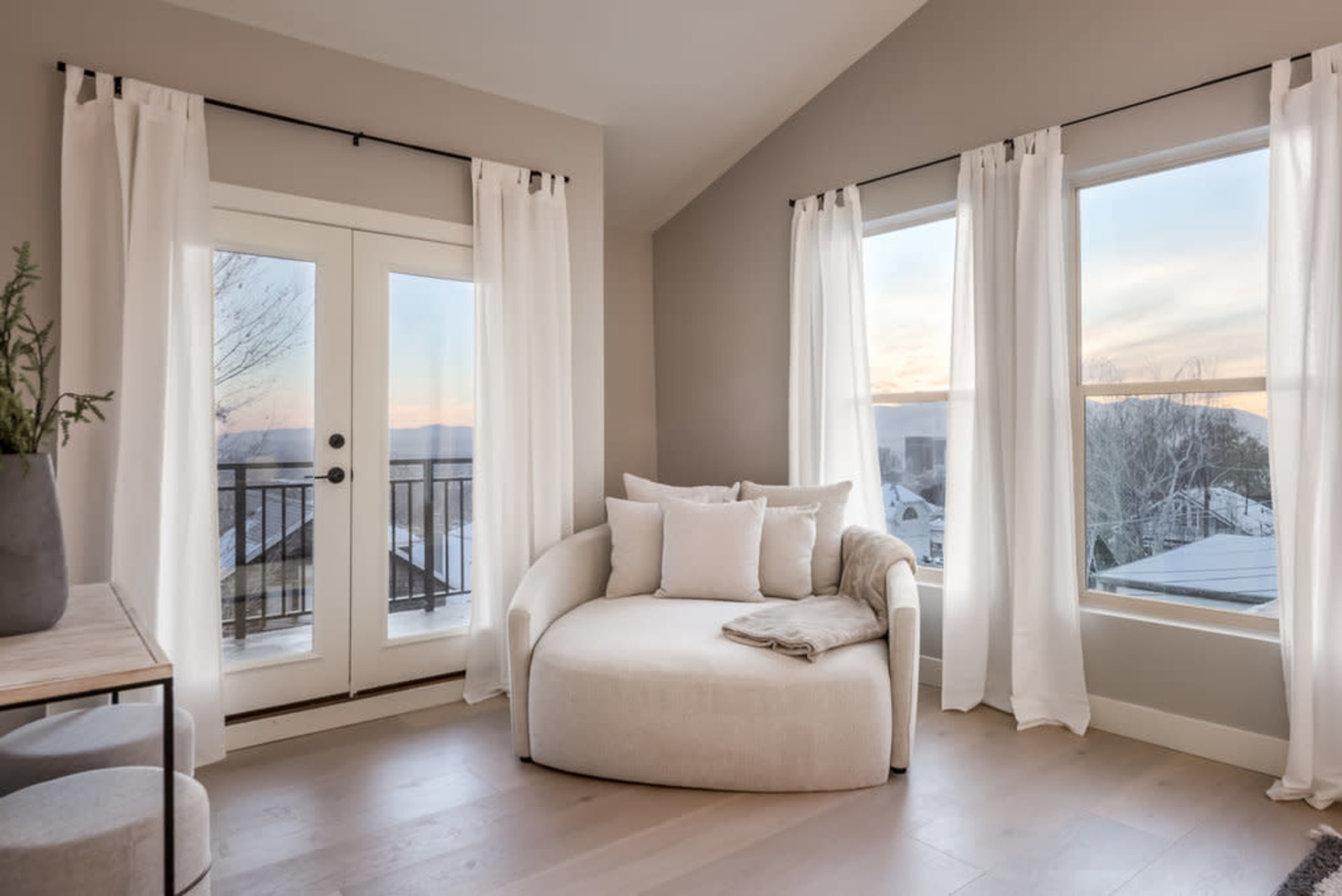 A cozy corner of a room features a curved sofa with pillows, large windows dressed in sheer white curtains, and a view of the outdoors.