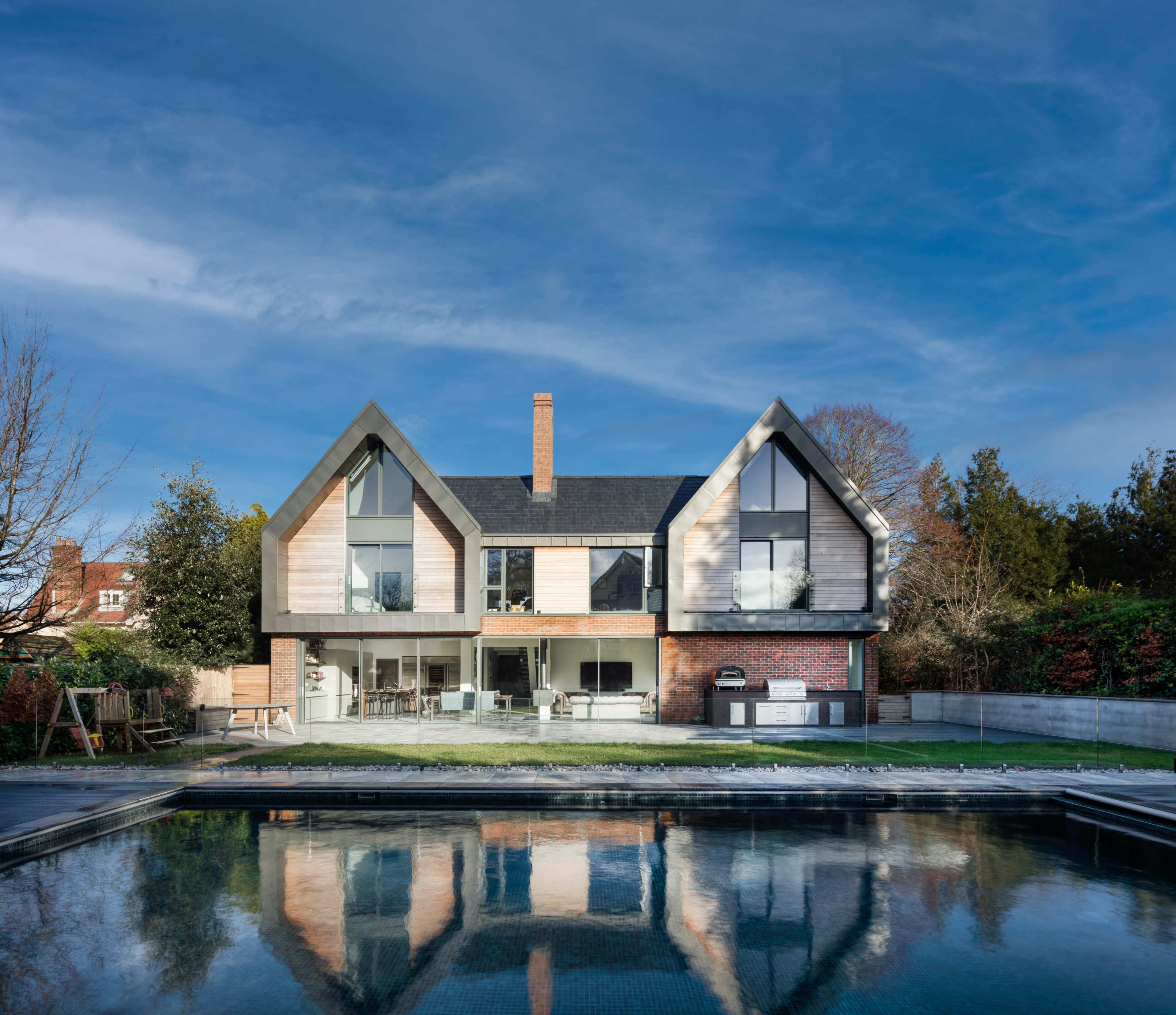 A modern two-story house features large windows and a combination of brick and wood siding, situated beside a reflective pool and surrounded by greenery.